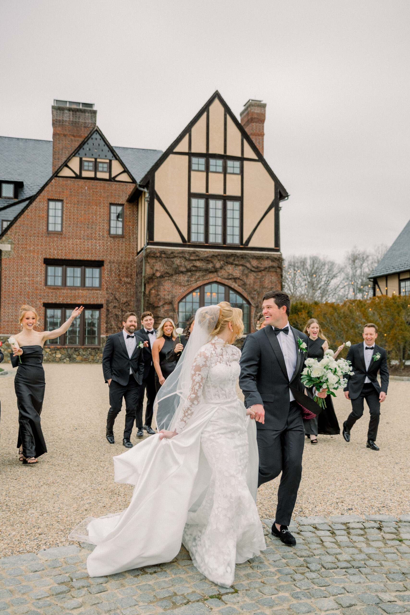 Wedding at Dover Hall in Manakin-Sabot, VA | Wedding Photographer in Richmond | bride in white floral gown and flowing veil holding hands with groom in black tux as they walk in front of Dover Hall Estate, followed by bridesmaids and groomsmen in black dresses and suits.