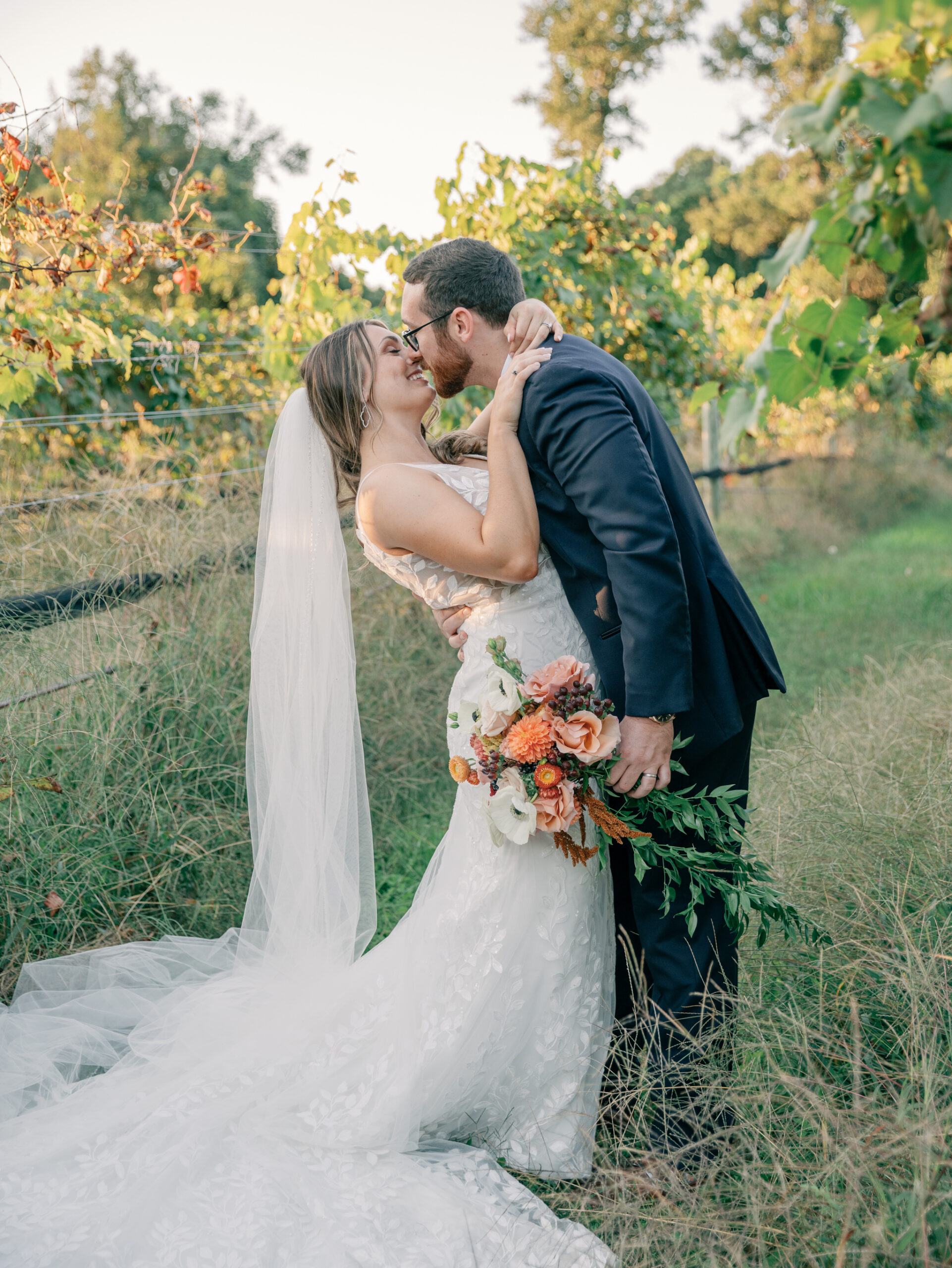 Wedding at Ashton Creek Vineyards Near Richmond, VA | Richmond Wedding Photographer | bride with long flowing veil and groom in navy suit kissing in the middle of a vineyard