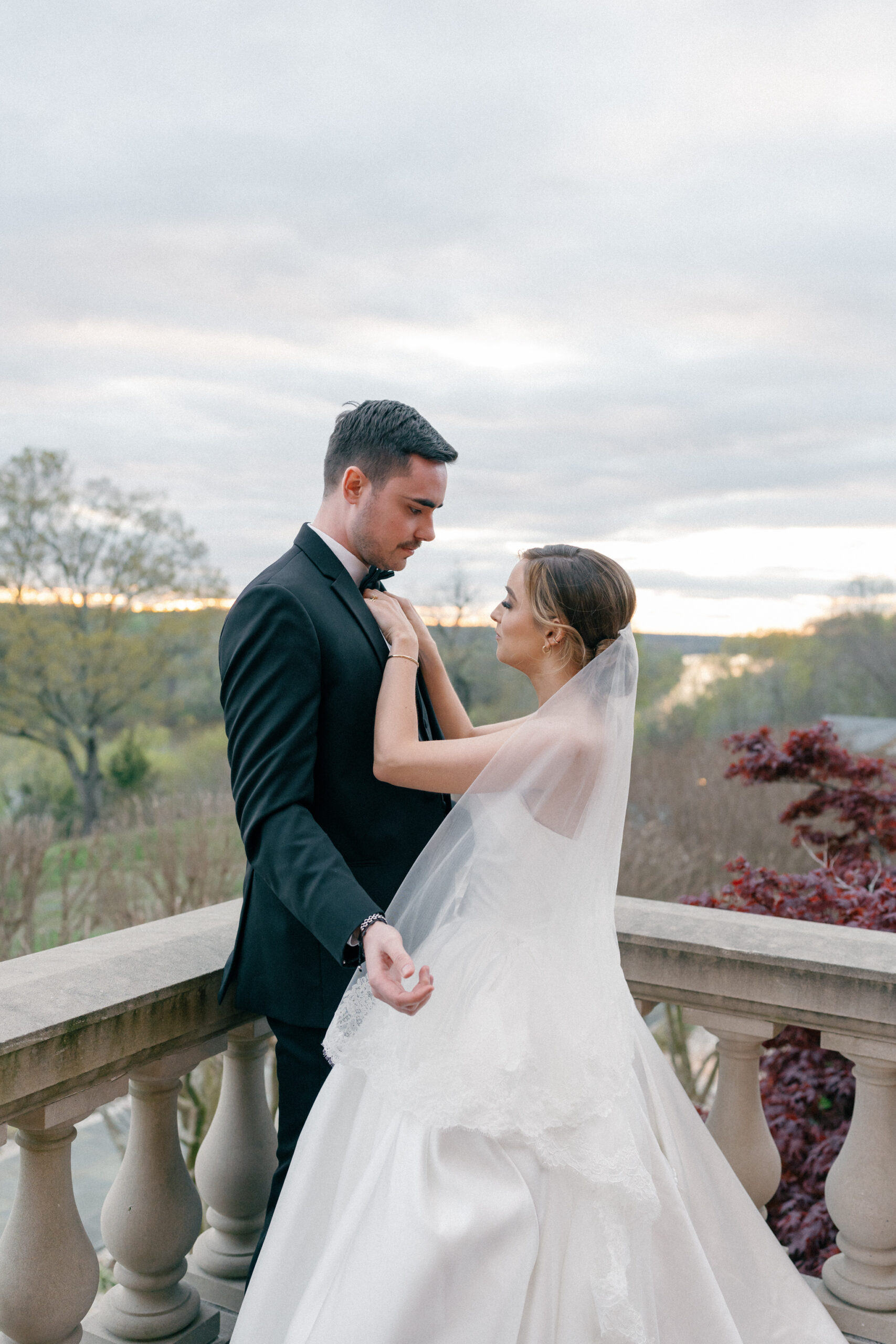 A romantic moment between a bride and groom on the terrace of The Estate at River Run. The bride gently adjusts her groom’s bow tie as they share an intimate gaze, framed by the soft glow of sunset and the rolling Virginia countryside in the background. Her delicate veil cascades down her gown, adding an ethereal touch to this timeless scene. Captured by Caitlin Wilcox Photography, a Richmond, VA film wedding photographer.