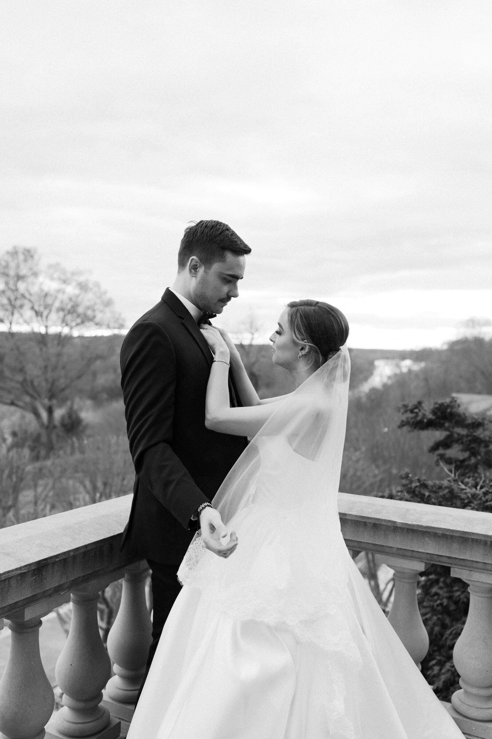 Estate at River Run Wedding in Richmond, VA | Richmond Wedding Photographer | black and white image of bride with veil blowing in the wind fixing her groom's bowtie on the balcony of the Estate at River Run