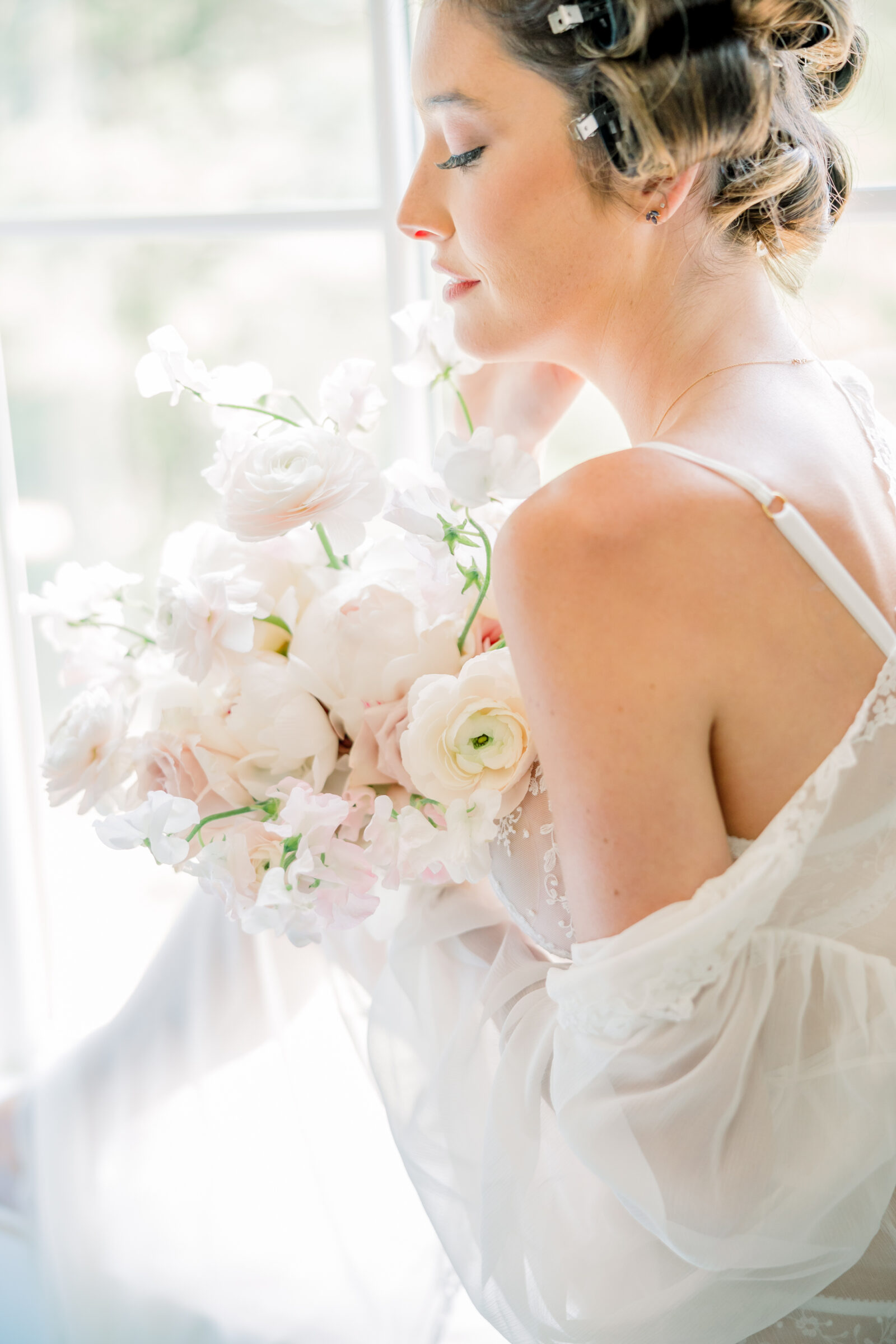 A softly lit, intimate portrait of a bride in a delicate, sheer gown, holding a bouquet of pale pink peonies, ranunculus, and sweet peas. Her hair is styled in soft curls, and she gazes serenely out of a window. This moment captures the elegance and timeless beauty of a fine art boudoir experience in Richmond, evoking a sense of romance and refinement.