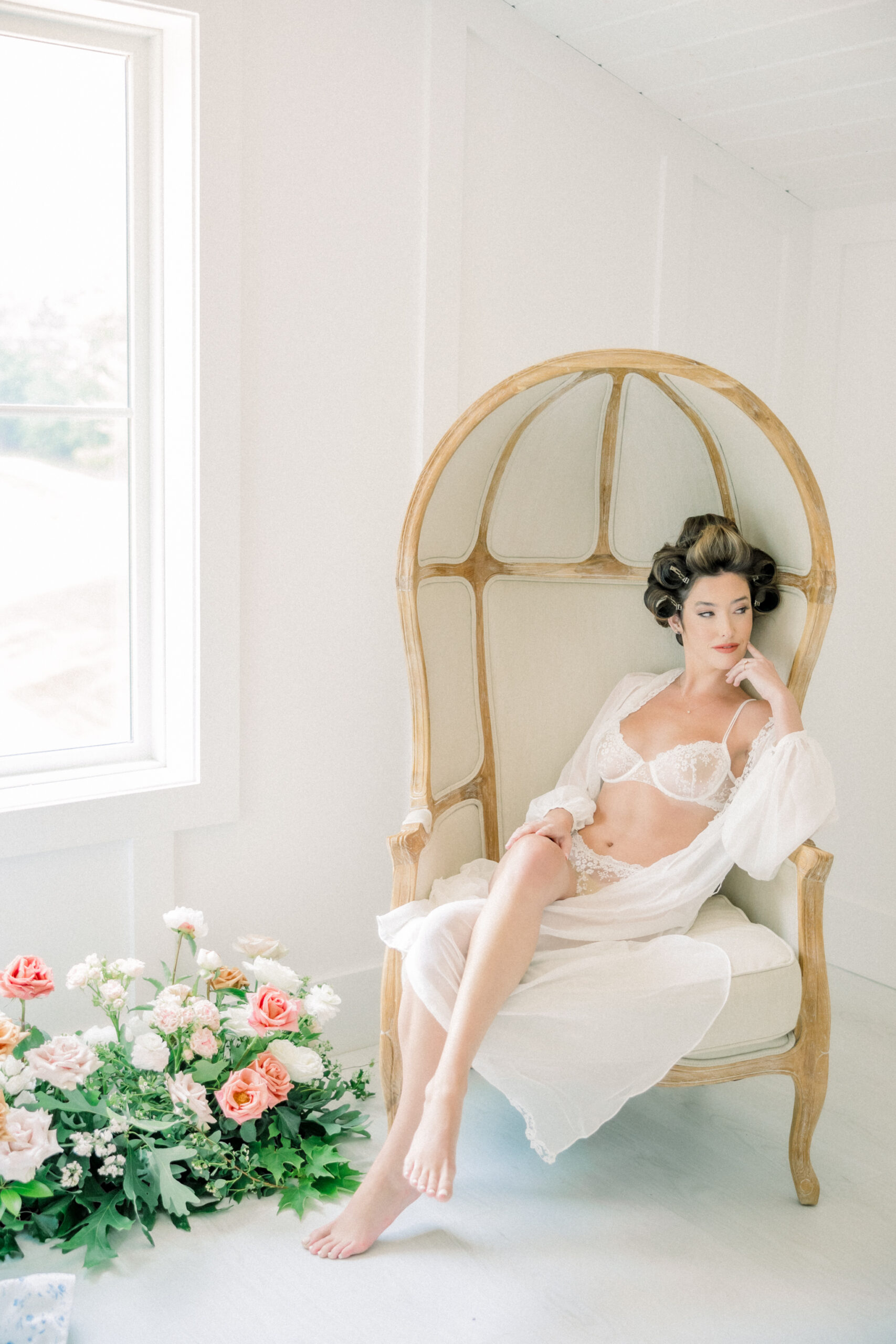 A model sitting gracefully in an elegant rattan chair, wearing a delicate white lace lingerie set and a sheer robe. Soft curls frame her face, and she rests her hand gently on her chin. In front of her, a lush arrangement of pink and white roses adds a romantic touch. The bright, airy room highlights the ethereal and timeless beauty of a fine art boudoir experience in Richmond.