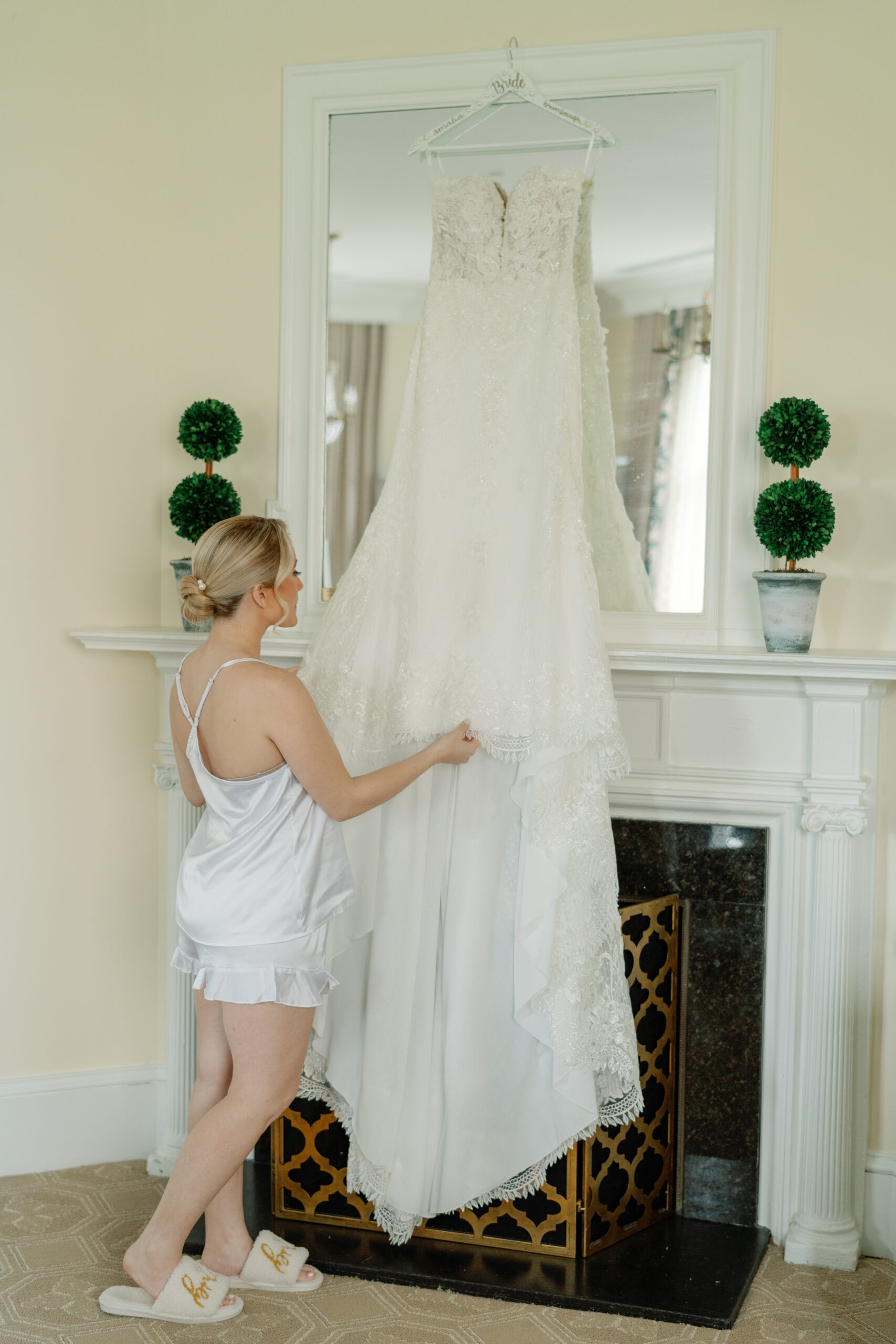 Wedding at the Jefferson Hotel in Richmond | bride in white satin pajamas and white bridal slippers looks at her wedding dress hanging above a fireplace in one of the suites of the Jefferson Hotel.