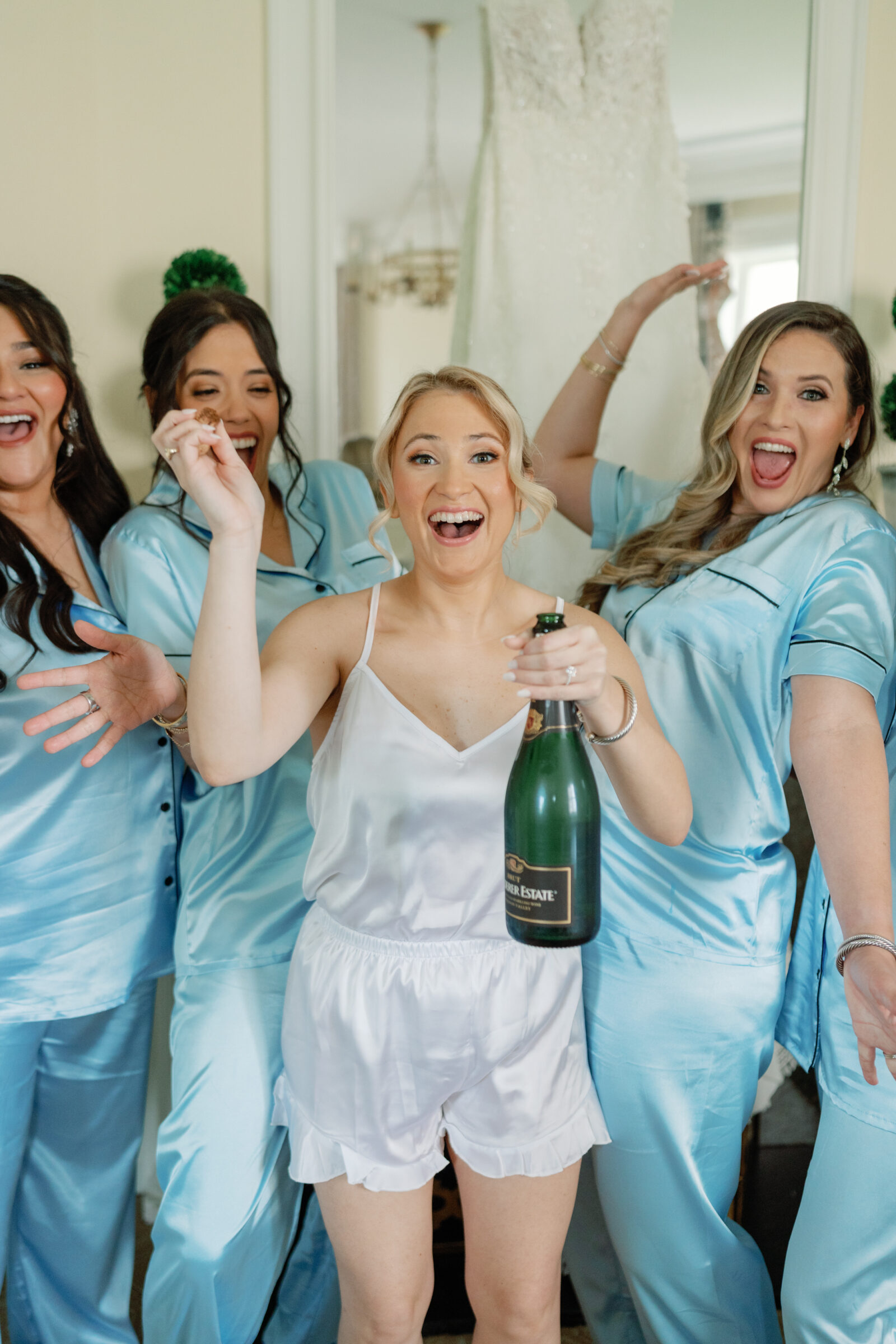 Jefferson Hotel Wedding in Richmond, VA | bride in white satin pajamas pops a bottle of champagne in her suite of the Jefferson Hotel, one of the best wedding venues in downtown Richmond. She is surrounded by her bridesmaids in light blue satin pajamas celebrating.