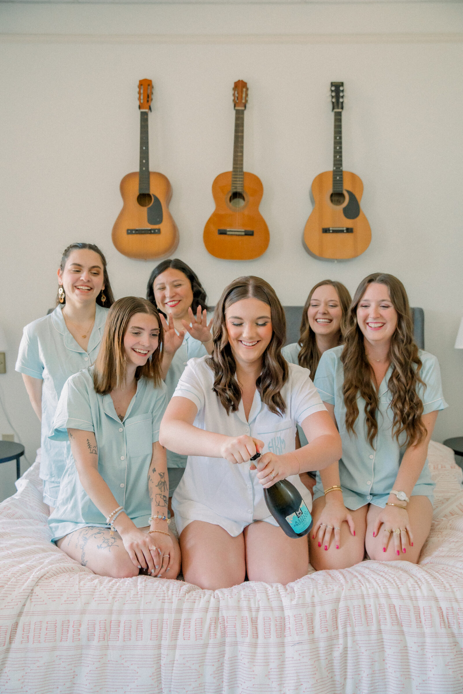 Lynchburg Wedding Photographer | bride in white pajamas opening a bottle of champagne while sitting on a bed surrounded by her bridesmaids in blue pajamas