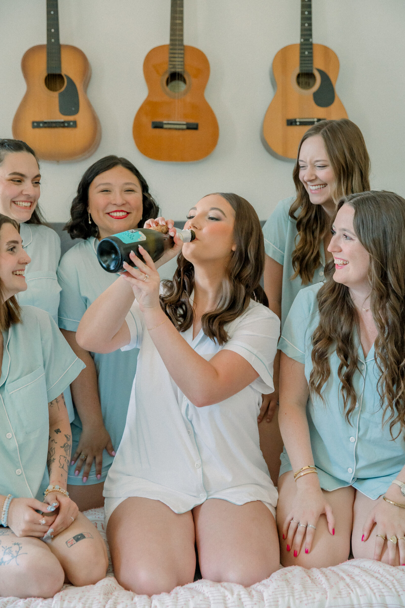 Lynchburg VA Wedding Photographer | bride in white pajamas drinks champagne out of the bottle while sitting on a bed surrounded by bridesmaids in blue pajamas