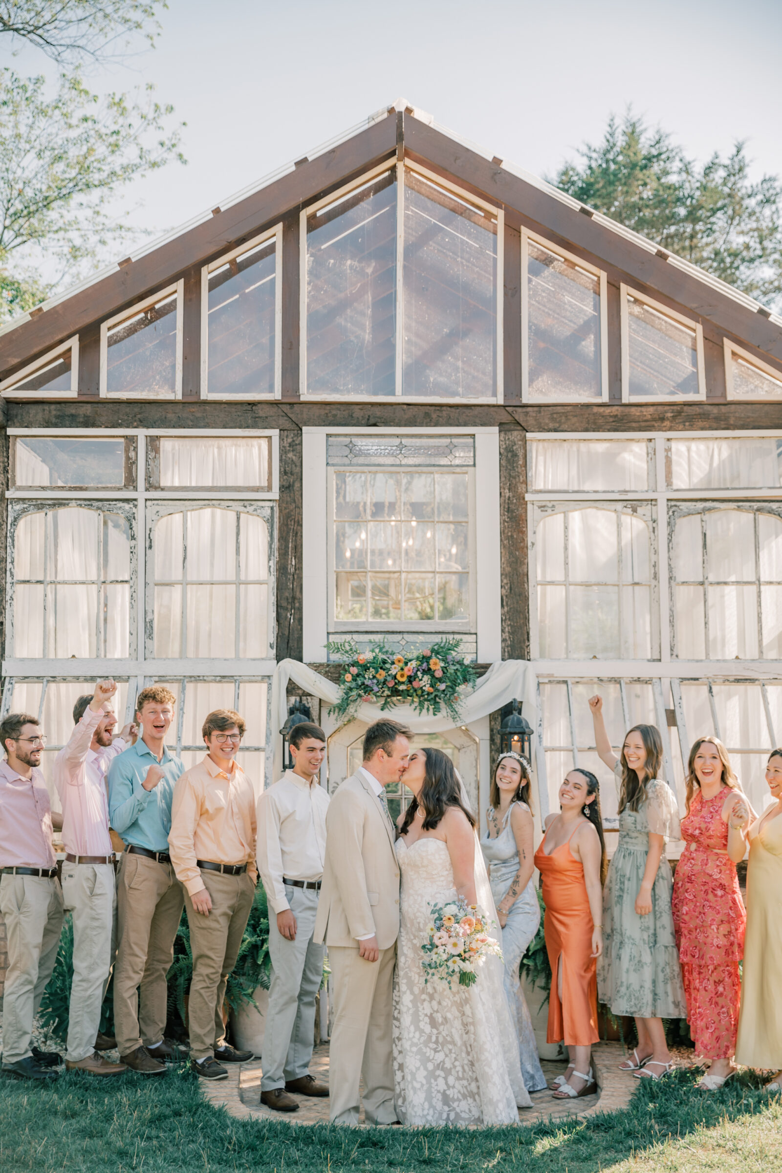 Three Oaks Manor Wedding in Madison Heights, VA | Virginia Wedding Photographer | bride and groom share a kiss in front of the greenhouse at Three Oaks Manor while wedding party celebrates them