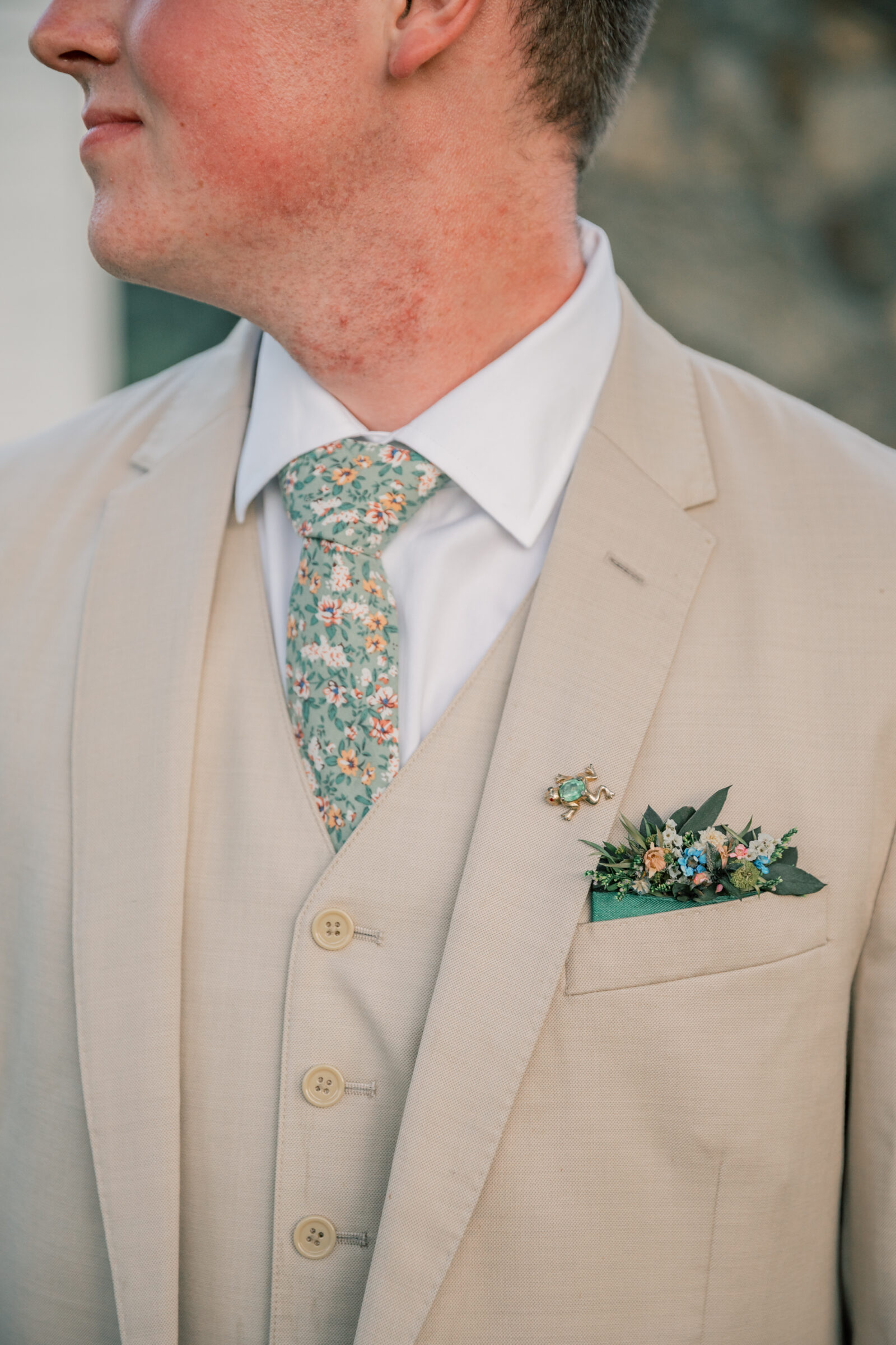 Three Oaks Manor Wedding Photographer | closeup of groom in tan suit and floral tie wears a green frog pin on his lapel