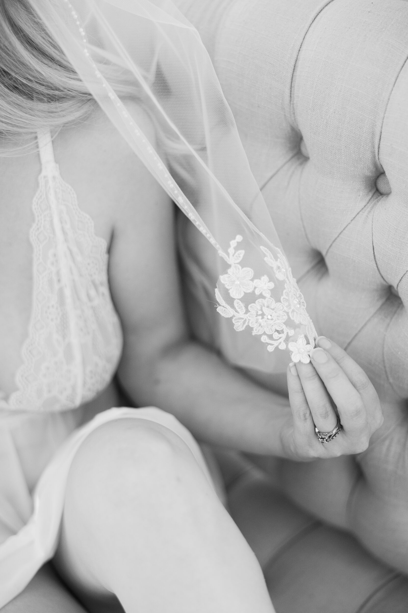 A black-and-white close-up of a woman gently holding her delicate lace veil, adorned with intricate floral embroidery. She is seated on a tufted chair, dressed in a soft, white lingerie set. The image exudes a romantic, bridal-inspired boudoir aesthetic, highlighting the grace and elegance of the Richmond VA boudoir experience.