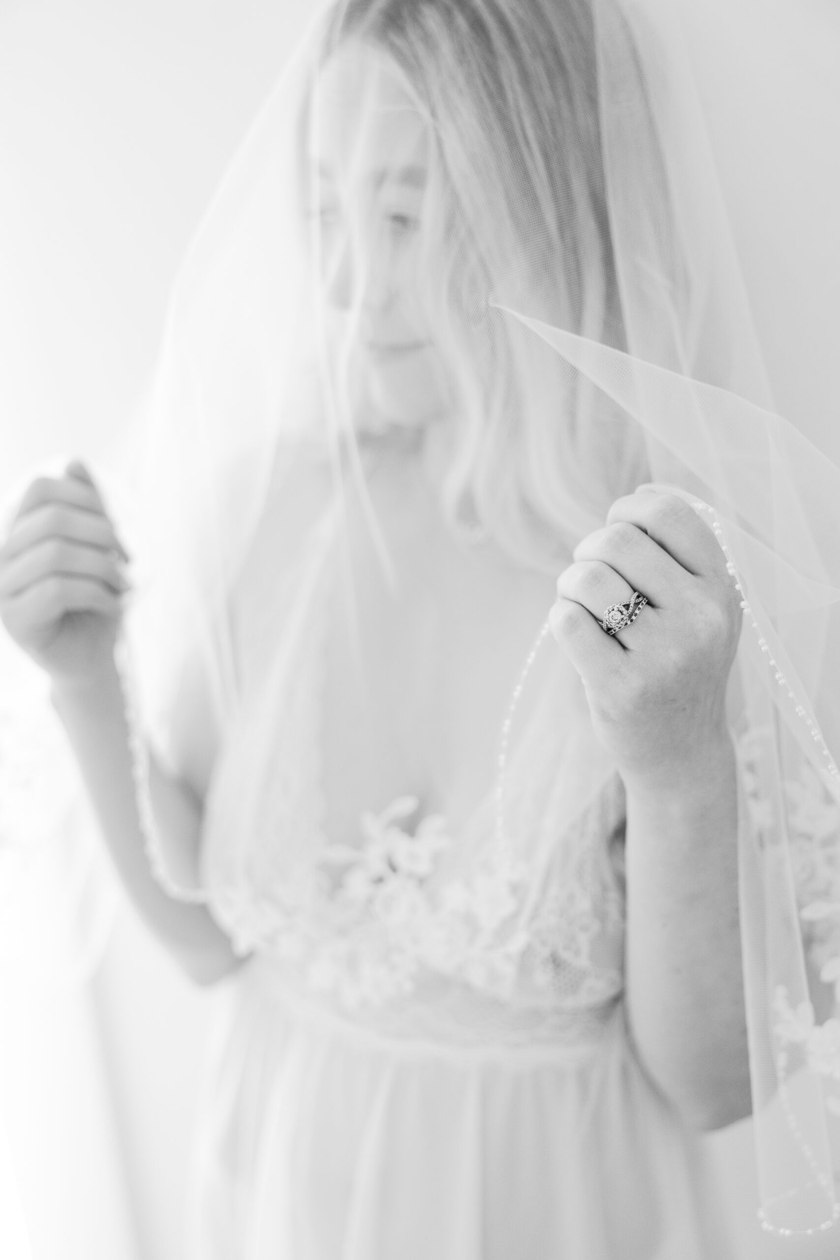 A soft, romantic boudoir shot of a woman gently holding her veil, with a delicate lace lingerie set. The veil drapes over her, creating a light, ethereal effect. She gazes softly into the distance, with her wedding rings elegantly displayed. The composition emphasizes an intimate, classic moment, with soft lighting that enhances the romantic atmosphere of the image. The focus is on her serene expression and the fine details of the lace and veil, evoking a sense of timeless beauty and elegance.