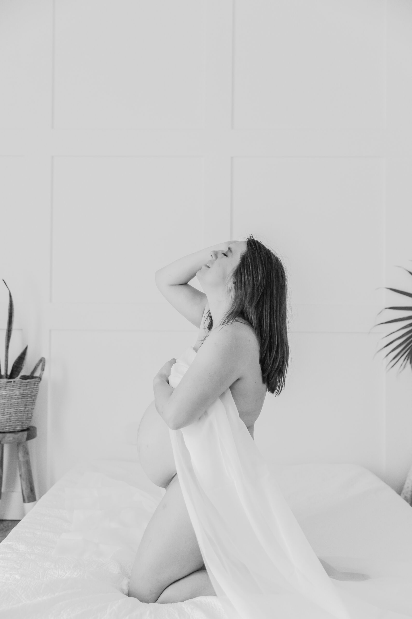 A maternity boudoir session featuring a pregnant woman draped in a white fabric, posing gracefully on a bed. The black and white image captures her holding the fabric to her chest, with her head tilted back and a soft, intimate expression. The minimalist background and soft lighting highlight her natural beauty and the tenderness of the moment.