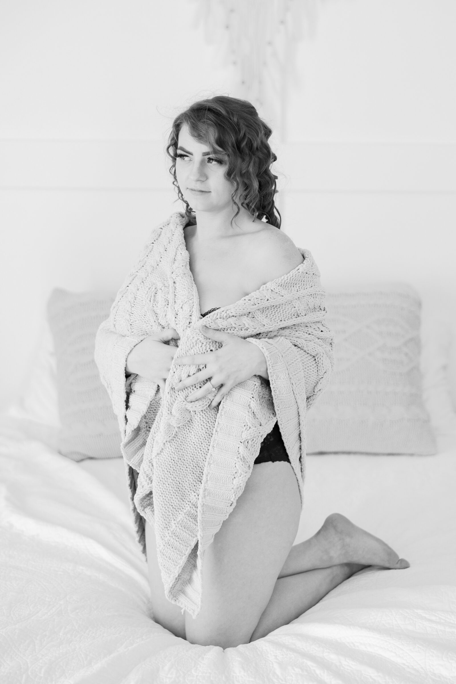 A black-and-white boudoir photograph of a woman kneeling on a bed, draped in a cozy knitted blanket. She gazes softly ahead with a relaxed, contemplative expression, showcasing her black lace lingerie. The soft, neutral tones of the image create a peaceful, intimate mood that reflects the elegance and confidence found in a Richmond VA boudoir experience.