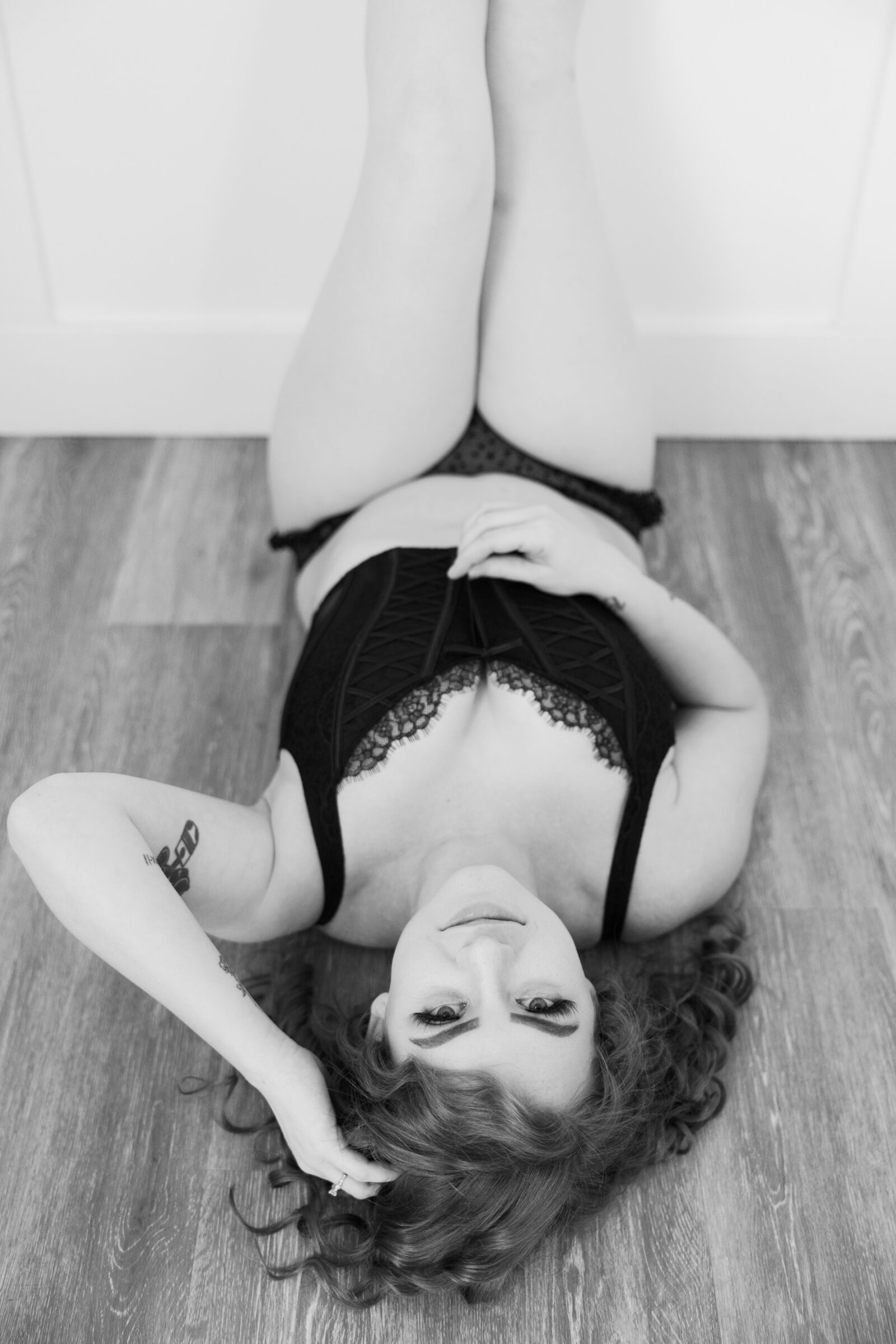 A black-and-white boudoir photograph of a woman lying on her back on a wooden floor, wearing black lingerie. She looks up at the camera with a relaxed and confident expression, her hand gently resting in her hair, highlighting her natural beauty and intimate pose.
