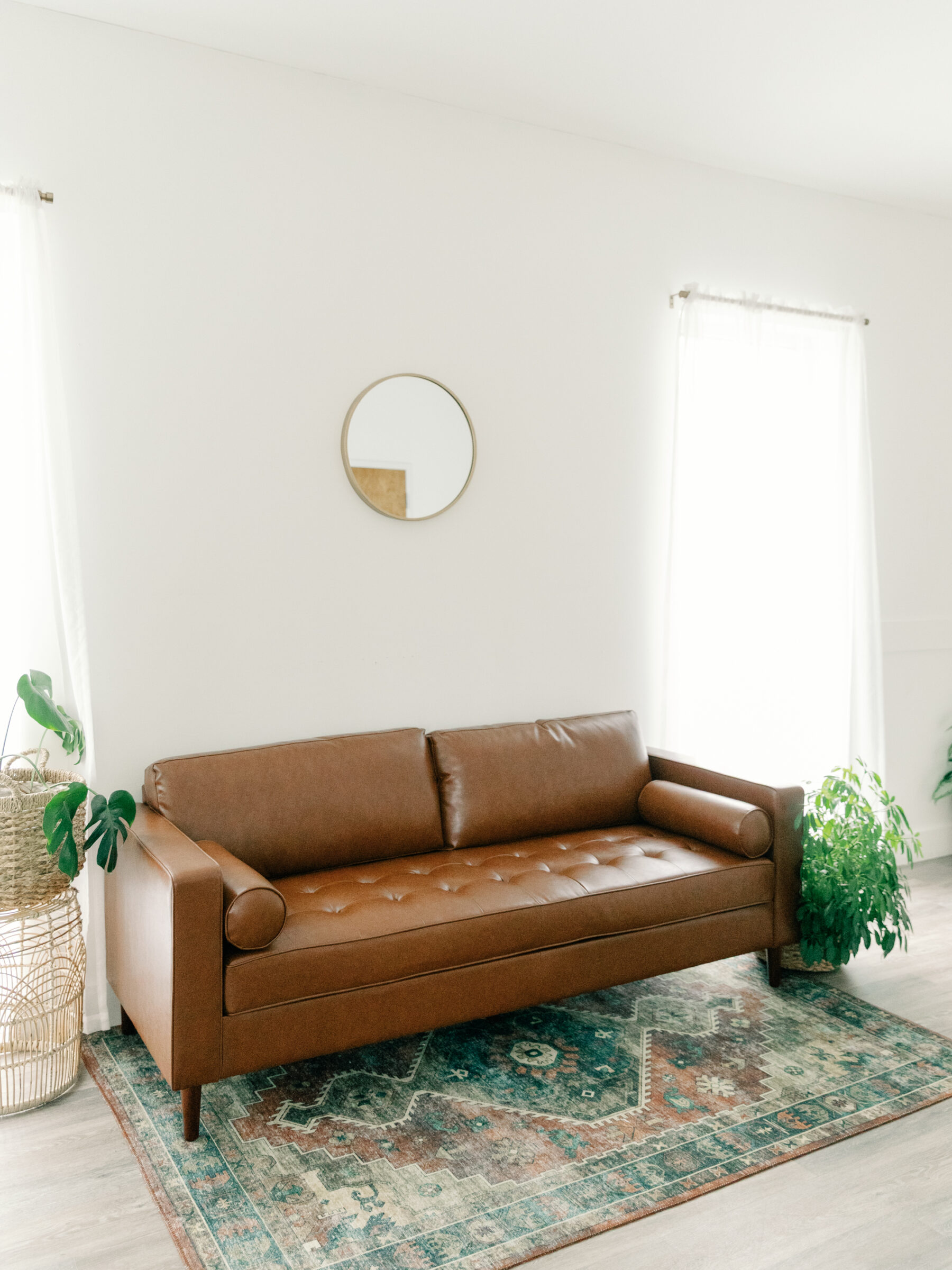 A cozy, minimalist corner of Carbone Studios in Ashland, VA, featuring a brown leather sofa, a patterned rug, and a mirror on the wall. Lush plants in woven baskets add a natural touch to the serene space.