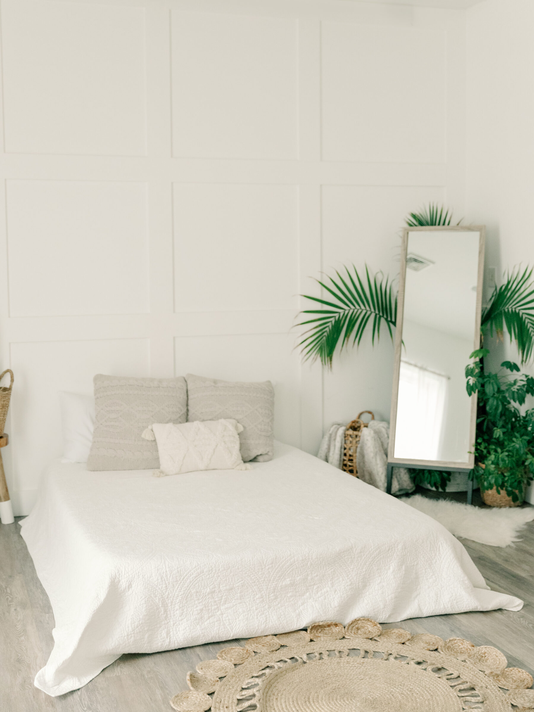 A cozy, minimalist boudoir photography studio in Ashland, VA near Richmond, featuring a soft white bed with textured pillows and a decorative rug. Lush plants and a large mirror create a serene, welcoming atmosphere for boudoir sessions at Carbone Studios, showcasing the studio's elegant and relaxing environment.