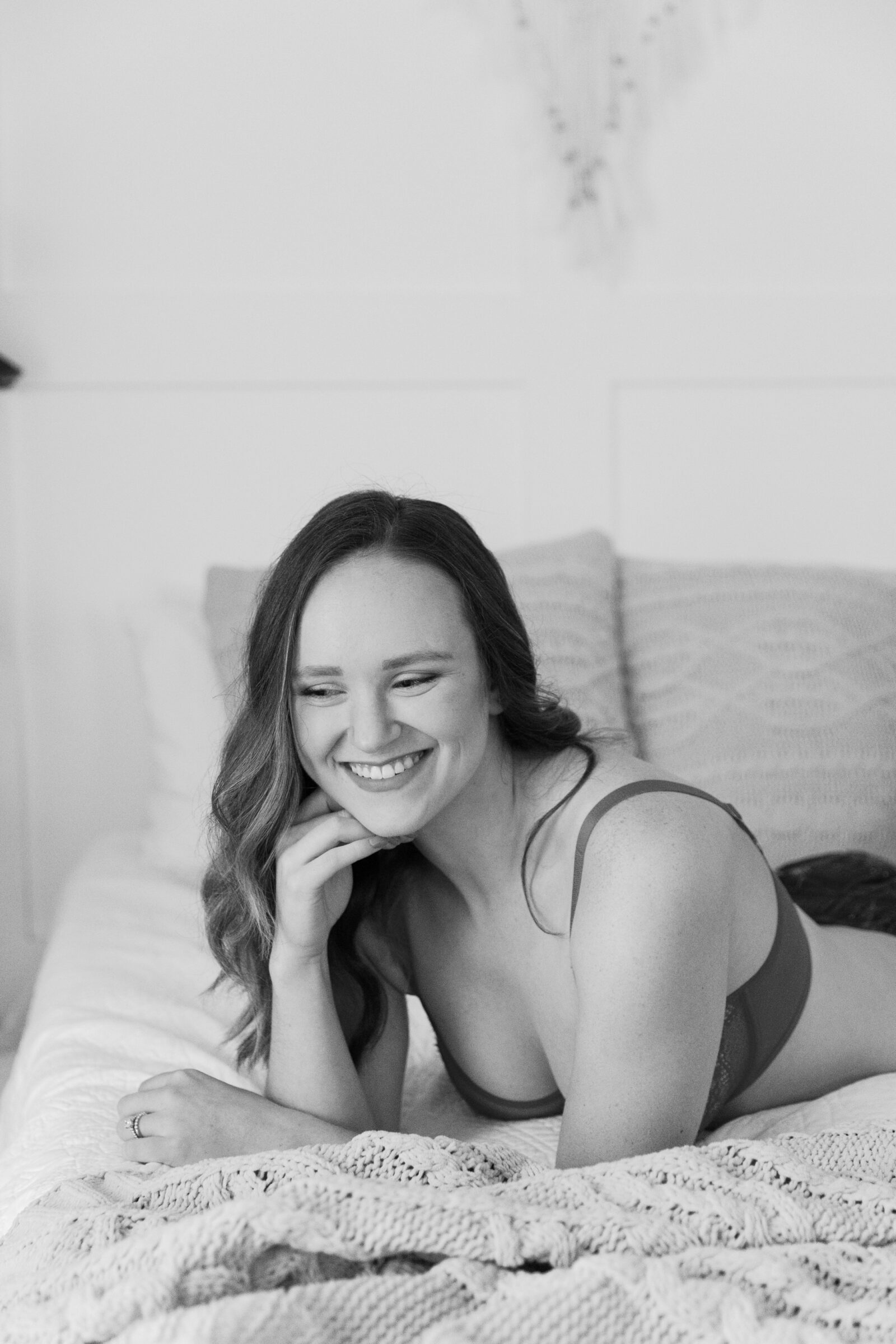 A woman smiles warmly while lying on a cozy bed at Carbone Studios in Ashland, VA. She is wearing a simple, elegant red lace bra and resting her chin on her hand. Soft, neutral-toned pillows and blankets create a relaxed and intimate atmosphere for this holiday boudoir session.