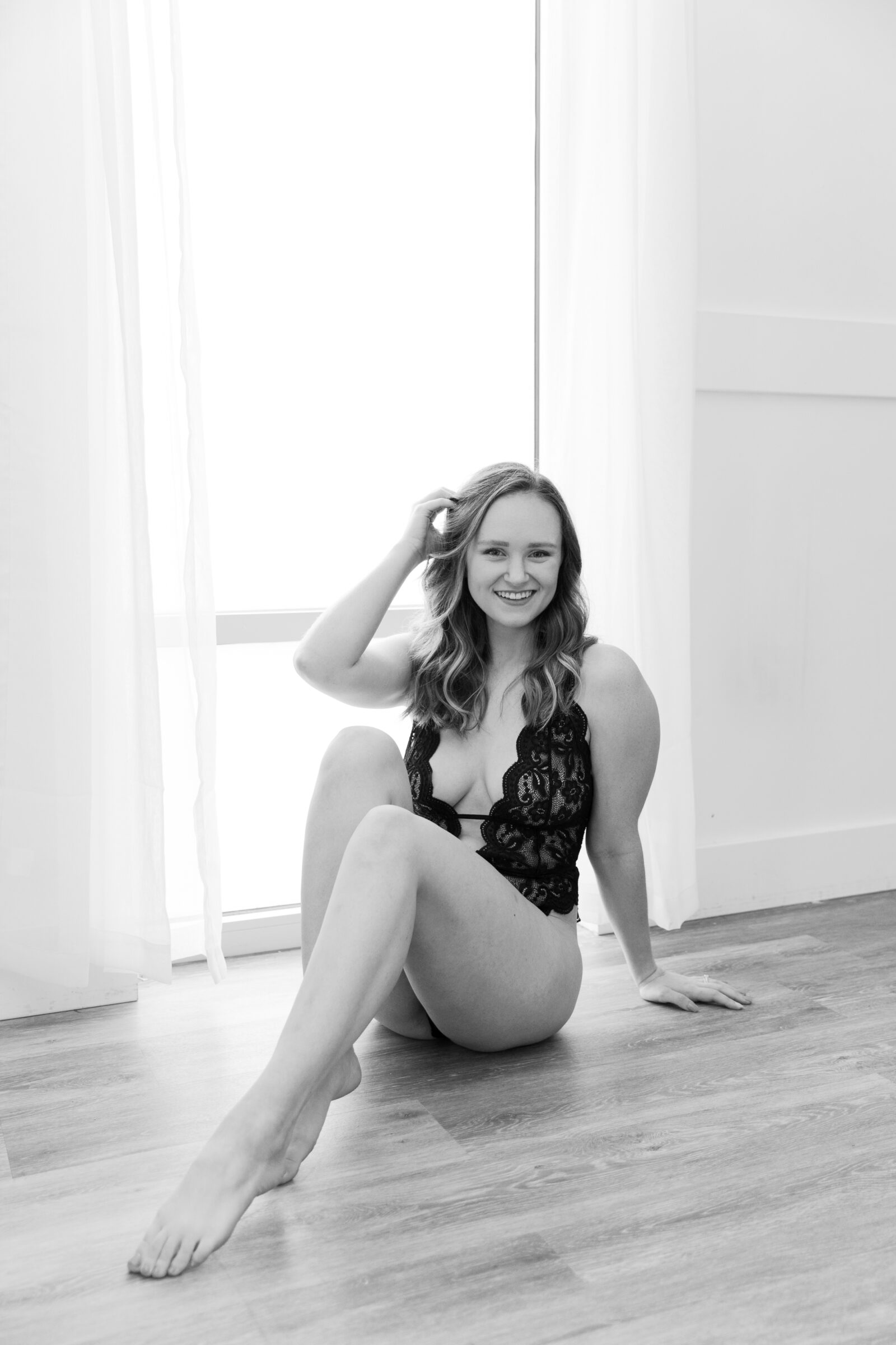 A black-and-white boudoir photograph of a woman seated on the floor, smiling warmly while playfully running her hand through her hair. She is wearing a black lace bodysuit and positioned in front of a window, with soft natural light casting a glow on her figure. The image conveys a relaxed, intimate, and confident vibe, with a sense of fun and ease.