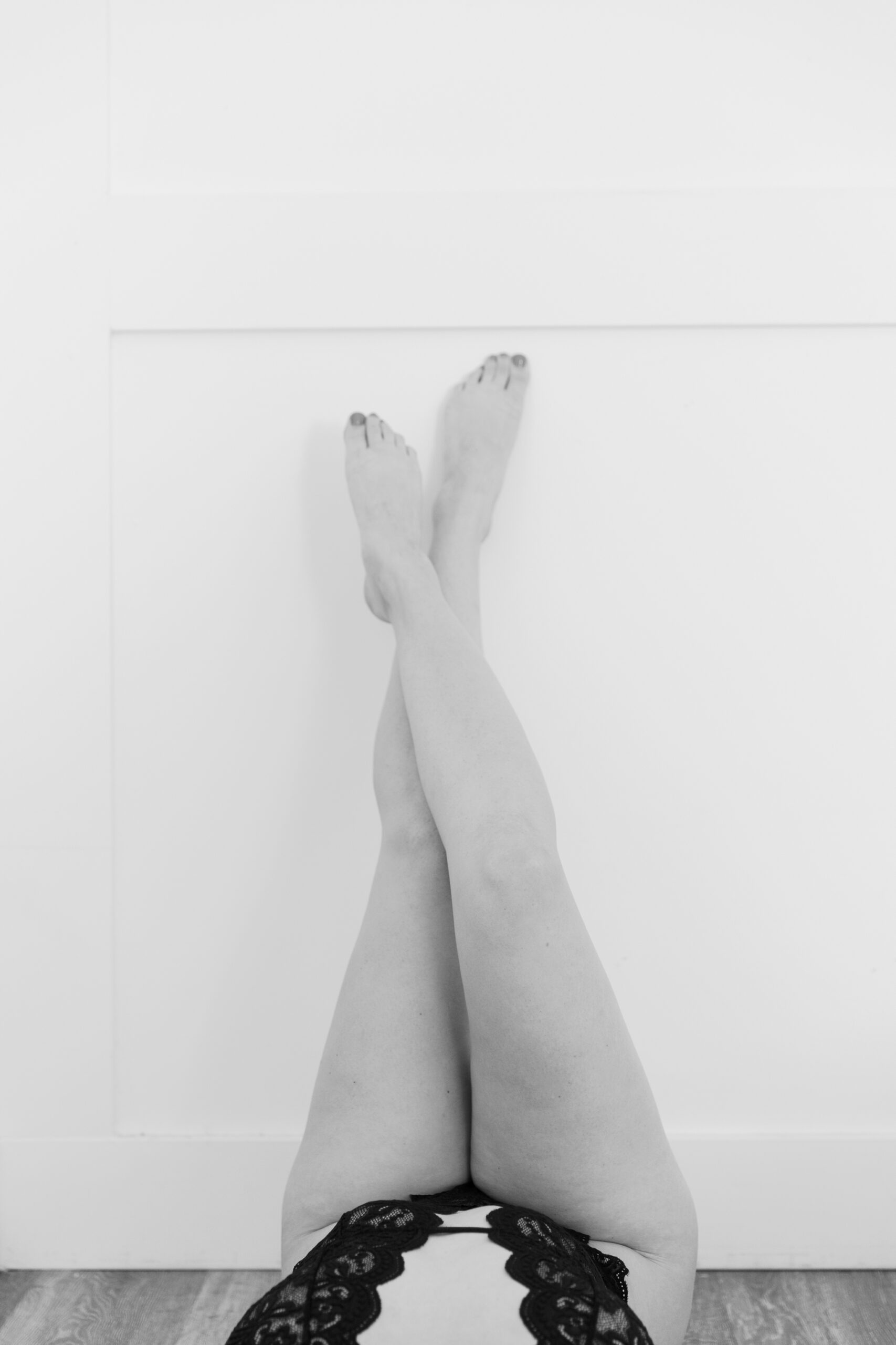 A black-and-white boudoir photograph of a woman’s legs crossed, showcasing her feet and the lace detailing of her black lingerie. The minimalist backdrop and simple floor create a serene, elegant atmosphere. This intimate shot is a beautiful example of the relaxed, confident vibe that comes with a Richmond VA boudoir experience.