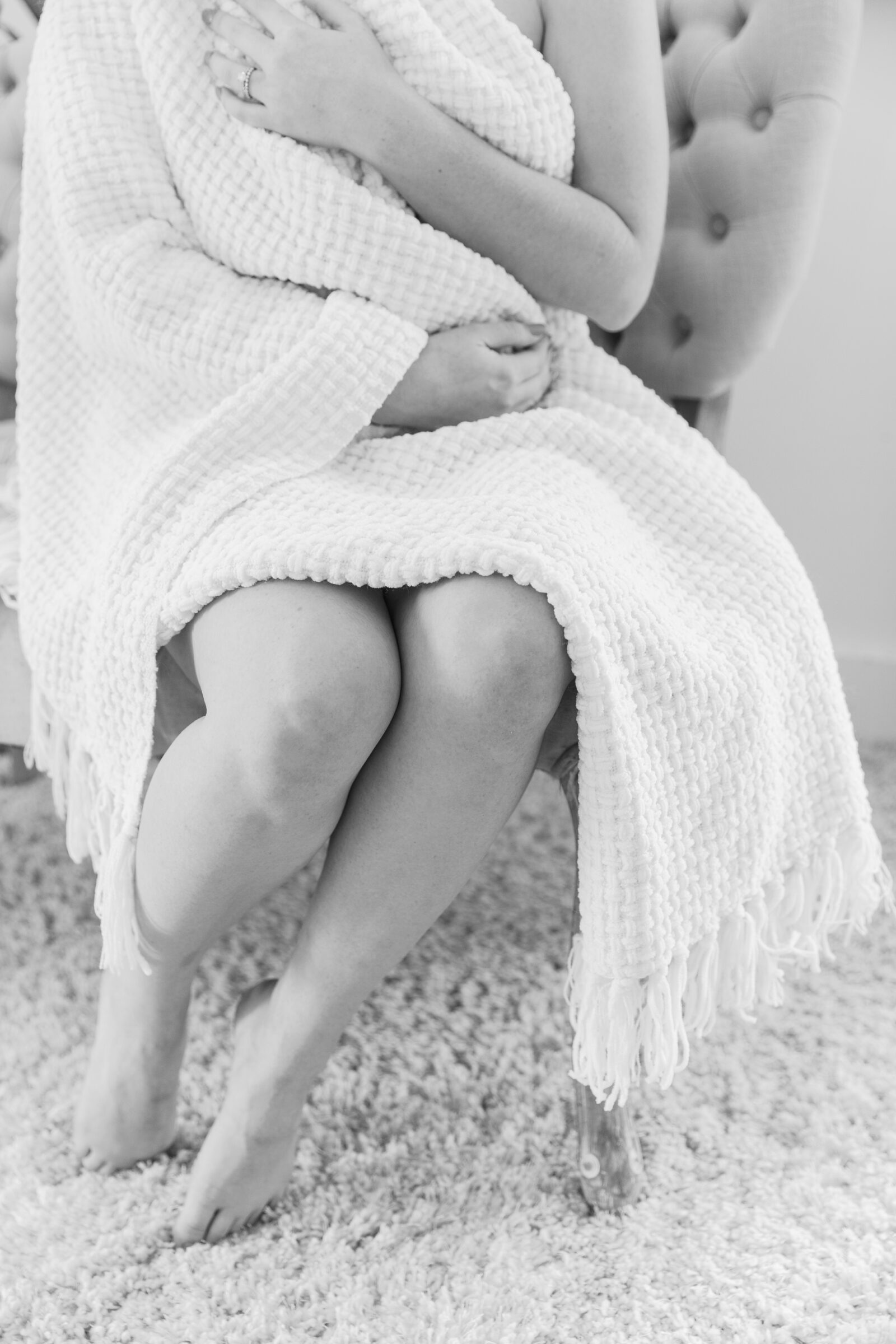 Richmond Boudoir Photographer | A soft and intimate boudoir shot of a woman seated in a plush chair, wrapped in a cozy white blanket. Her bare legs are crossed and resting on a plush rug. She gently holds the blanket with one hand while the other is placed across her chest, evoking a calm and relaxed atmosphere for her boudoir session at Carbone Studios in Ashland, VA.