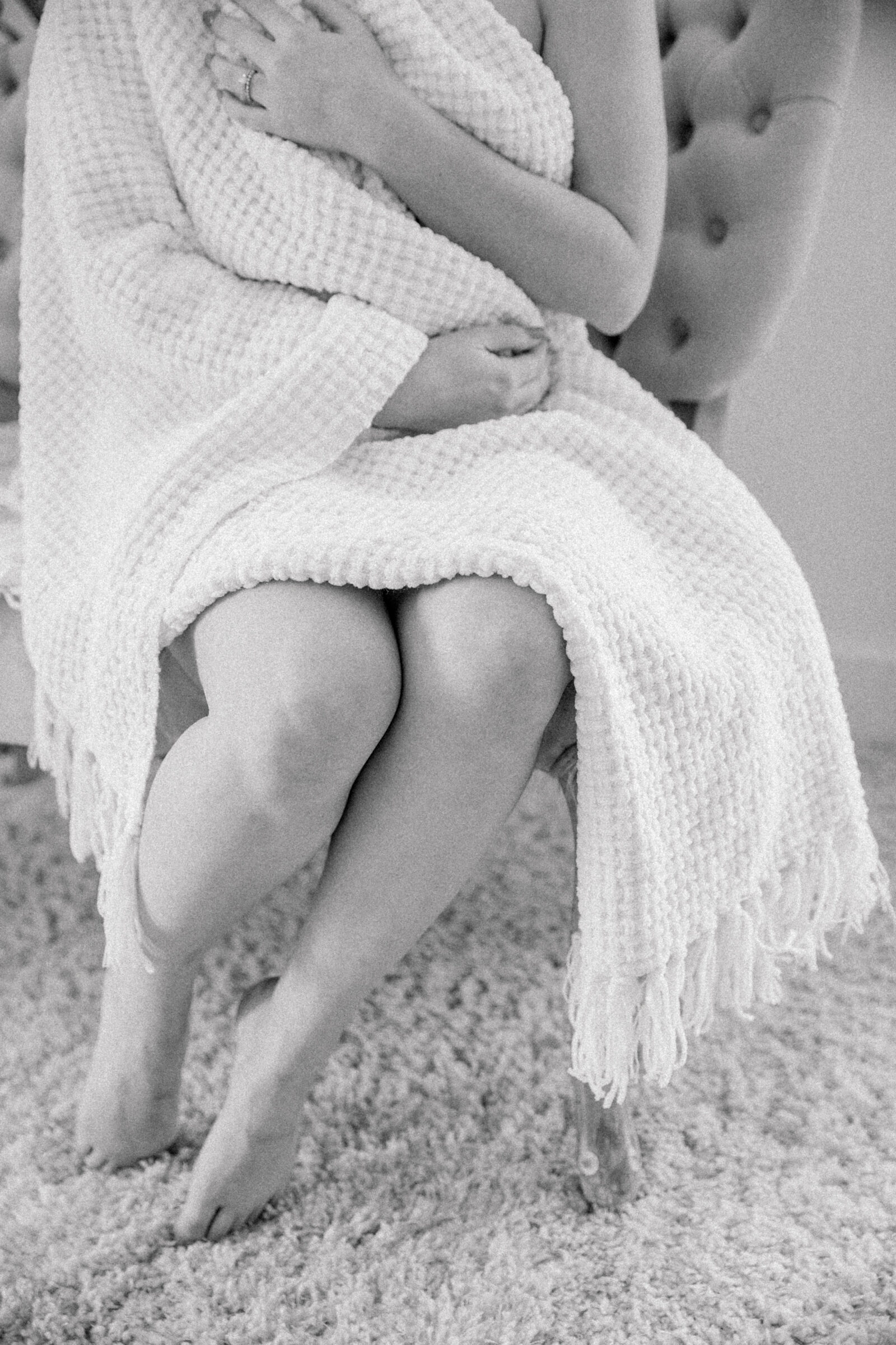 A woman sitting on a tufted chair, wrapped in a soft white towel, during a boudoir session with a top Richmond boudoir photographer. She holds the towel around her, exuding a relaxed and intimate vibe. The natural light from the window illuminates her skin, creating a serene and elegant atmosphere. The cozy, neutral-toned space adds to the comfortable, inviting feel of the shoot.