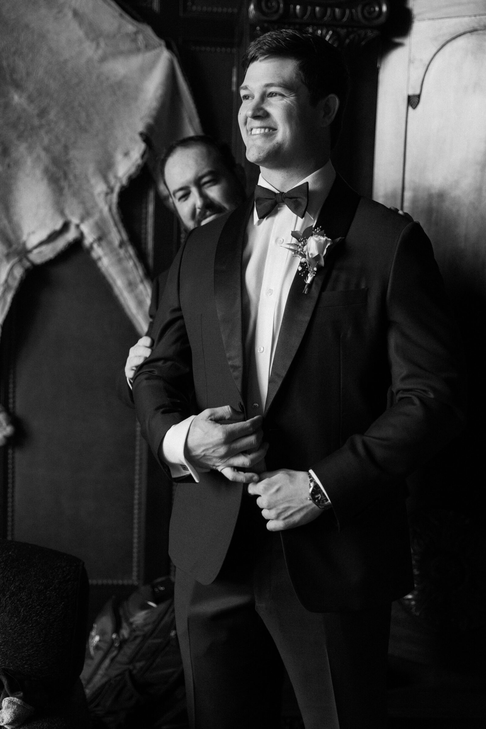 A groom smiling as his groomsman helps him adjust his tuxedo jacket in the groom's suite at Dover Hall Richmond VA.