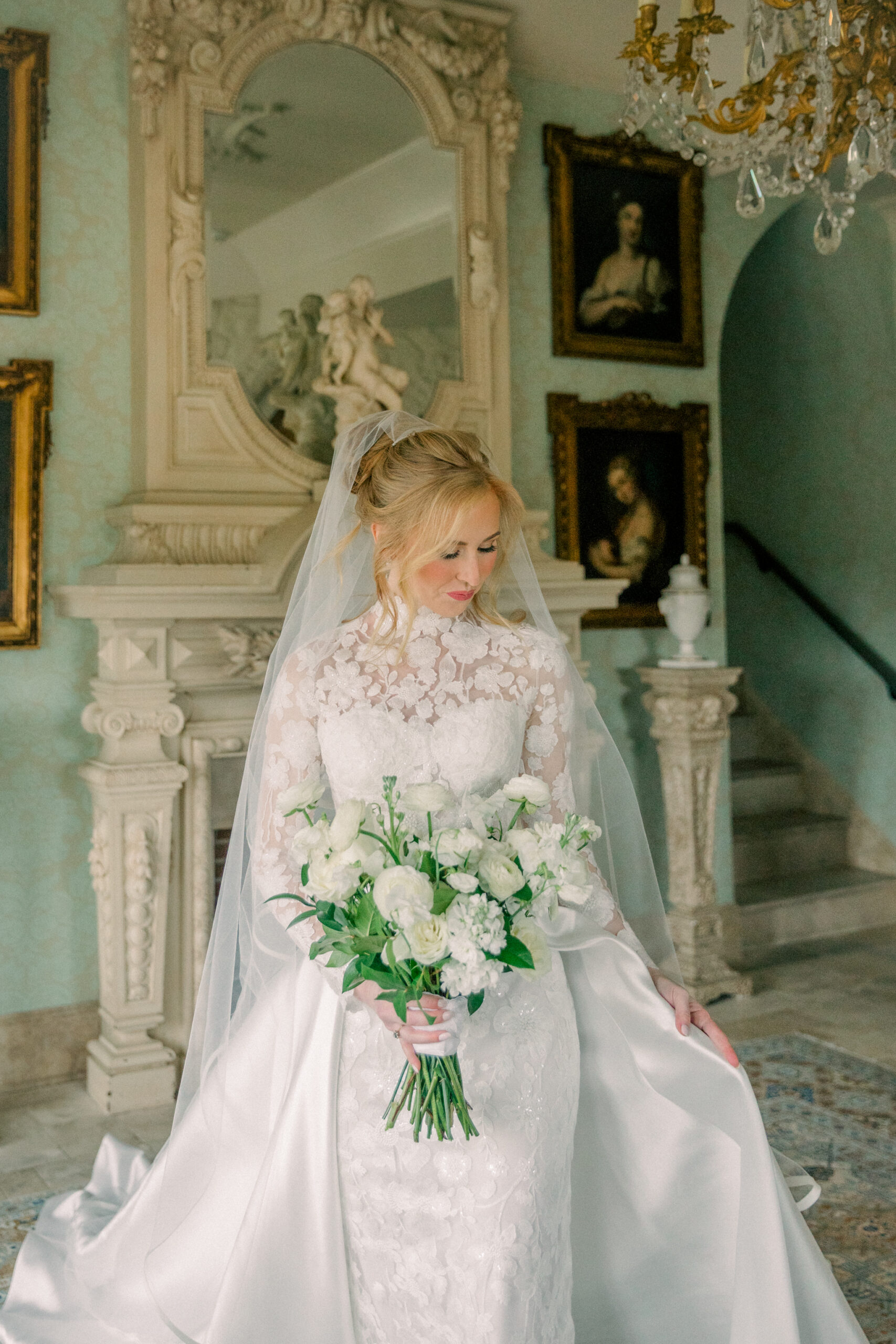 A bride in a lace wedding gown holding a bouquet of white flowers and standing in front of an ornate mirror inside Dover Hall Richmond VA, with a staircase visible in the background.