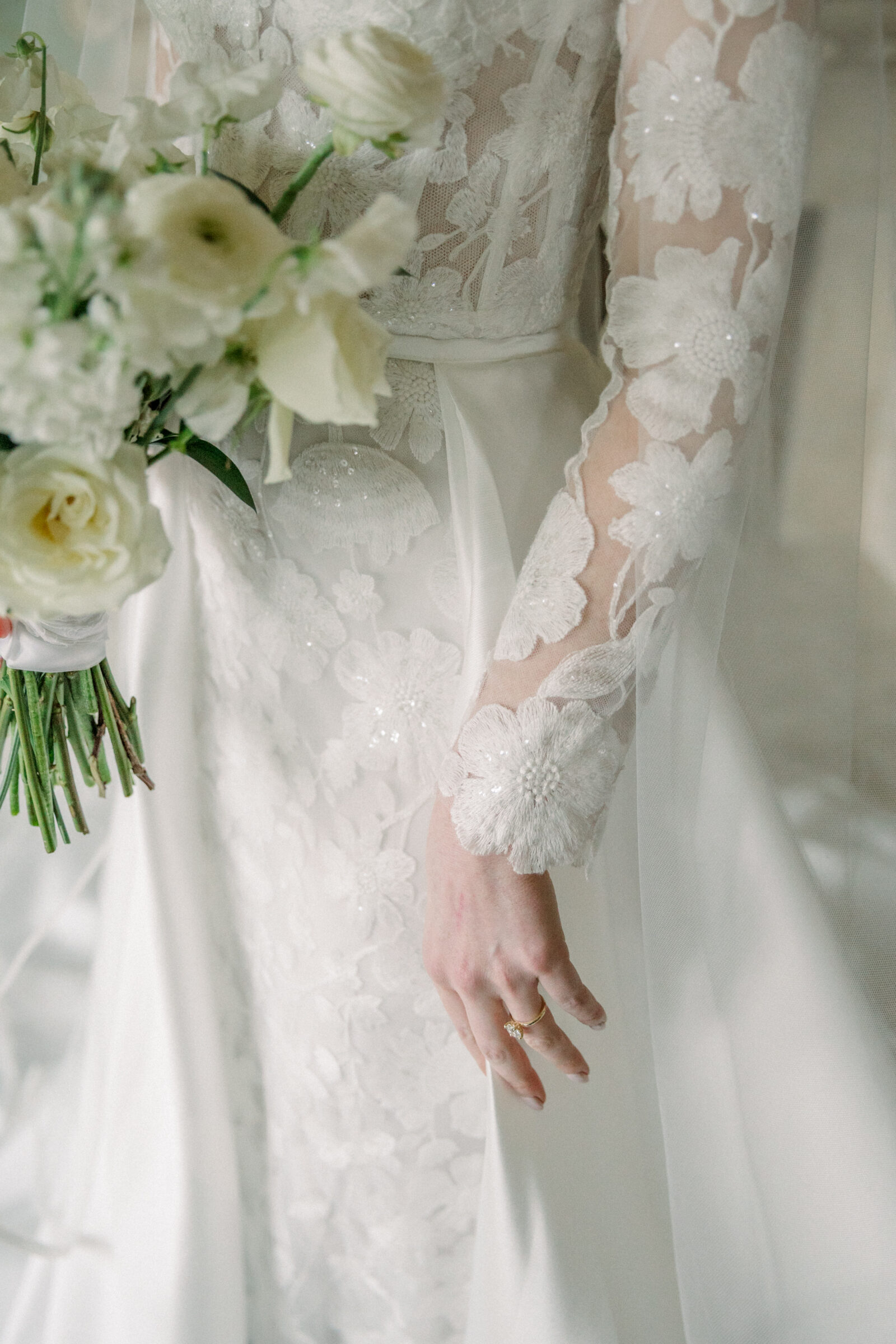 Close-up of a bride's hand holding a bouquet of white flowers, showcasing the detailed lace floral sleeve of her wedding dress at Dover Hall Richmond VA.