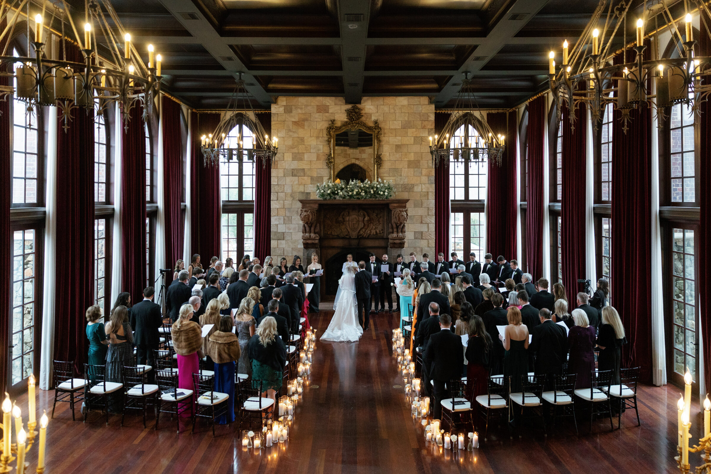 A stunning view of a wedding ceremony at Dover Hall Richmond VA, showcasing the grand, vaulted ceilings, and towering windows. The bride and groom stand at the altar surrounded by their guests, with candles lining the aisle, creating a romantic and elegant atmosphere.