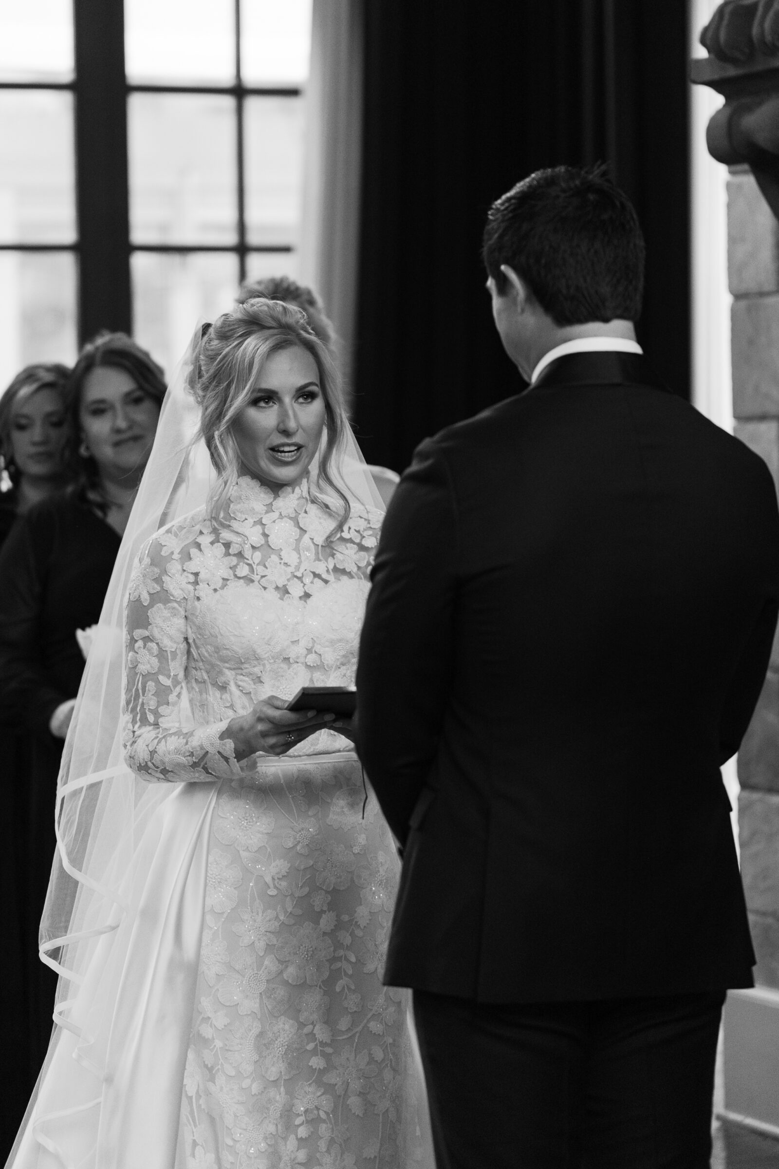 A stunning black-and-white photo of a bride at Dover Hall Richmond VA during her wedding ceremony, exchanging vows with her groom. She is wearing a lace long-sleeve wedding gown and a veil, holding a ring box while looking at her partner, surrounded by guests in the background.