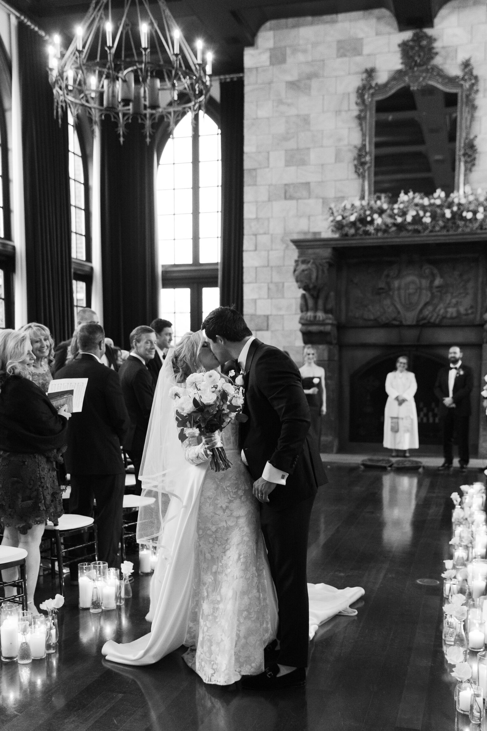 A beautiful black-and-white photo of a couple kissing after exchanging vows at their wedding at Dover Hall Richmond VA. The ceremony takes place in a grand hall with high windows, elegant stone walls, and soft candlelight, capturing a moment of joy and love.