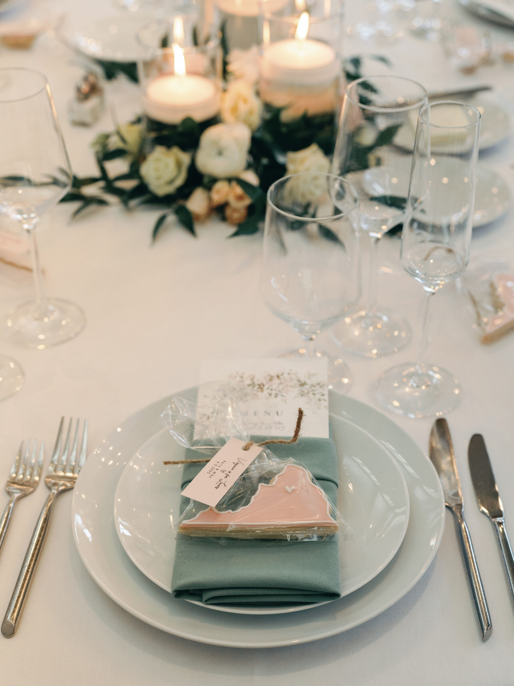 Elegant wedding reception table setting at King Family Vineyards featuring a mint green napkin, a personalized cookie favor, and a beautiful floral arrangement with lit candles, all arranged on a white tablecloth.
