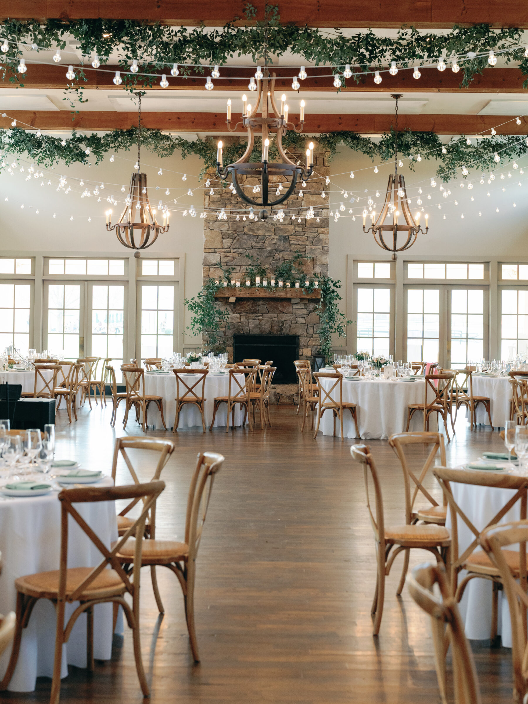 Interior view of the reception space at King Family Vineyards with rustic wooden beams, string lights, and elegant chandeliers. The space features round tables with white linens and wooden chairs, with a large stone fireplace as a focal point.