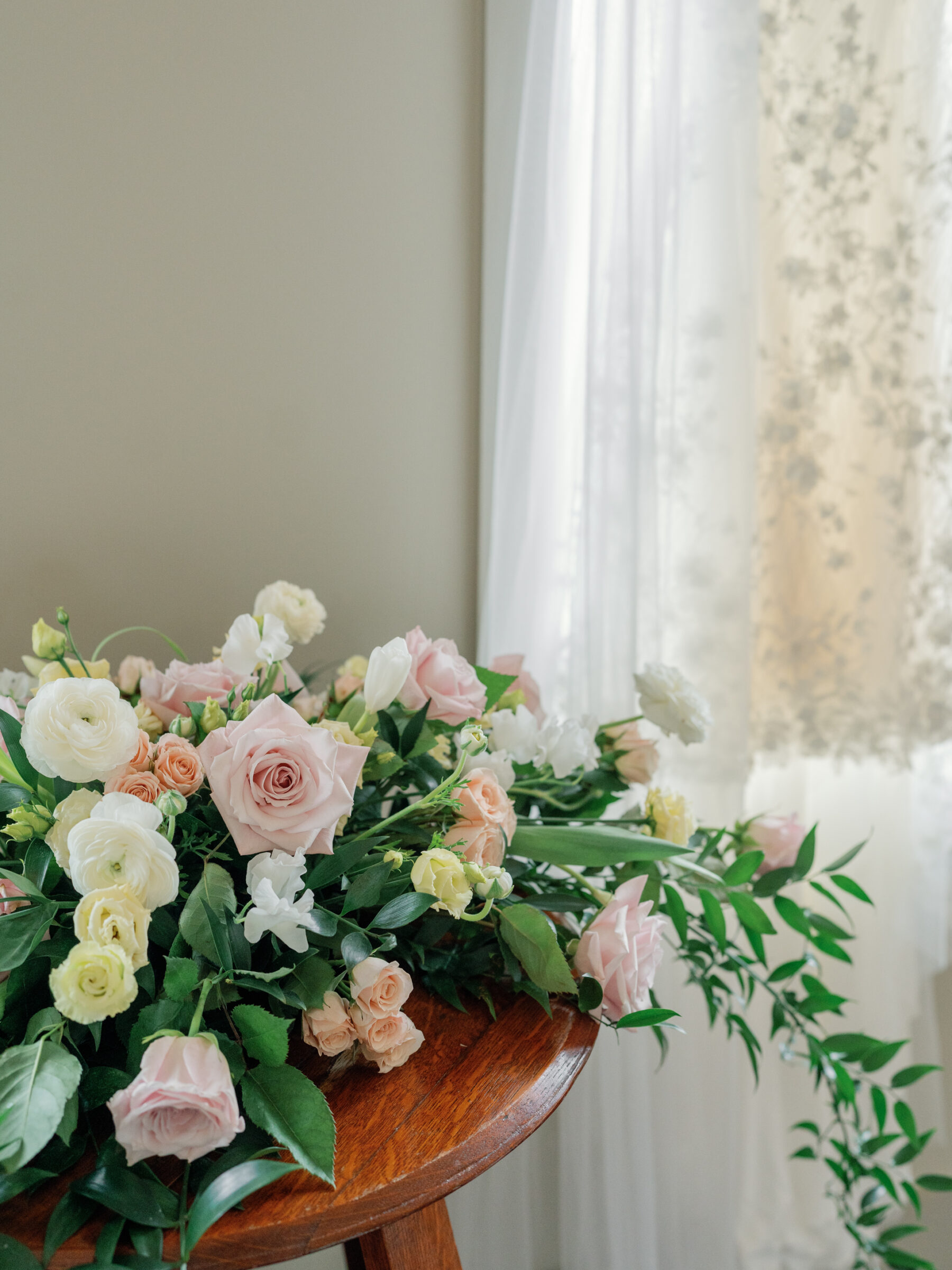 A beautiful bridal bouquet overflowing with soft pink roses, white and yellow ranunculus, and delicate greenery arranged on a wooden table, with sunlight gently filtering through a sheer curtain in the background.