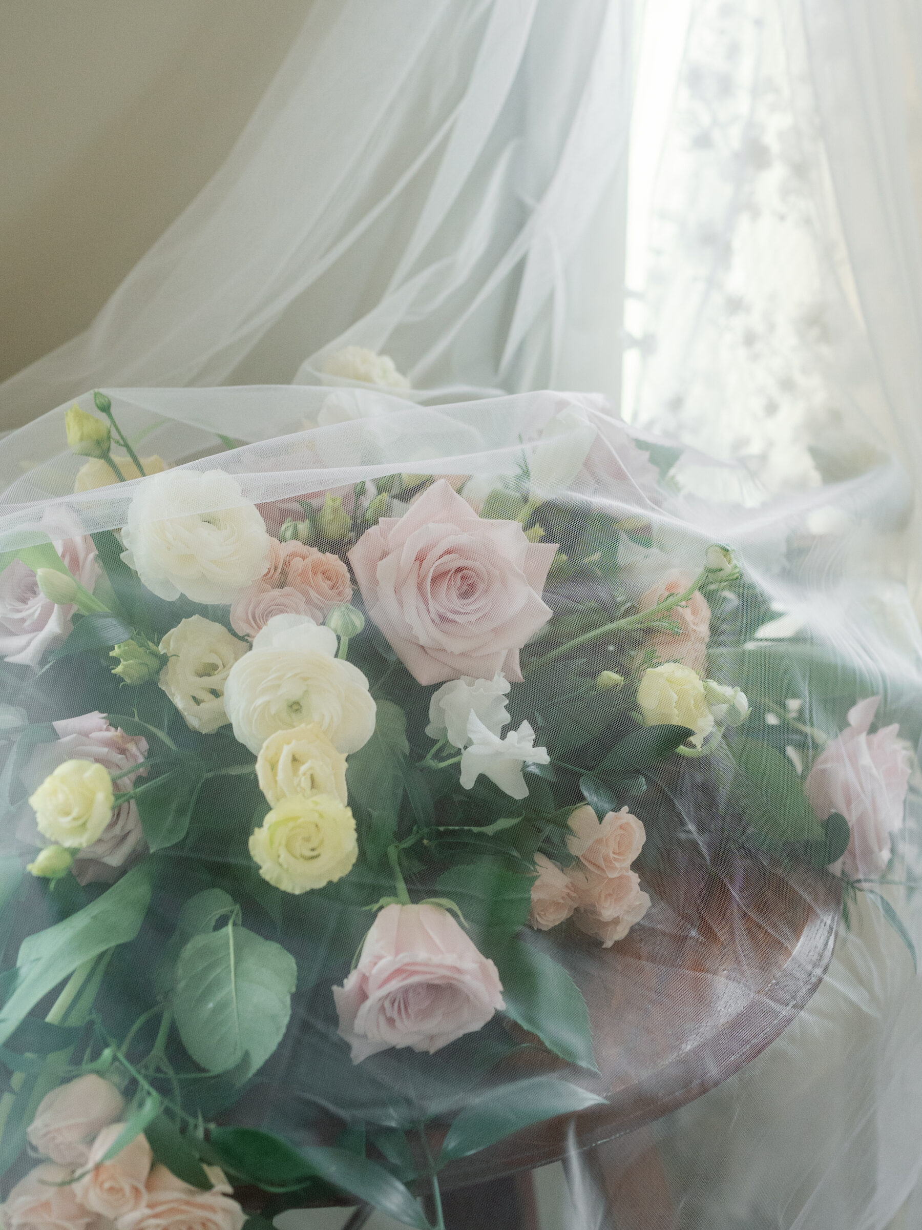 A soft veil drapes over a romantic bridal bouquet of pale pink roses, yellow ranunculus, and delicate white flowers, elegantly arranged on a wooden table with gentle natural light in the background.