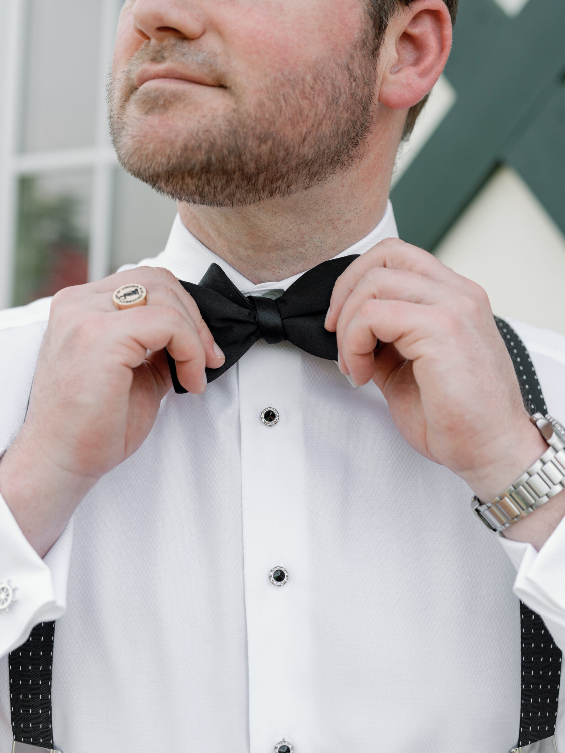 A groom adjusts his black bow tie with one hand while holding it in place with his other hand. He wears a white dress shirt with cufflinks, black suspenders, and a silver watch, and the focus is on his face and bow tie.