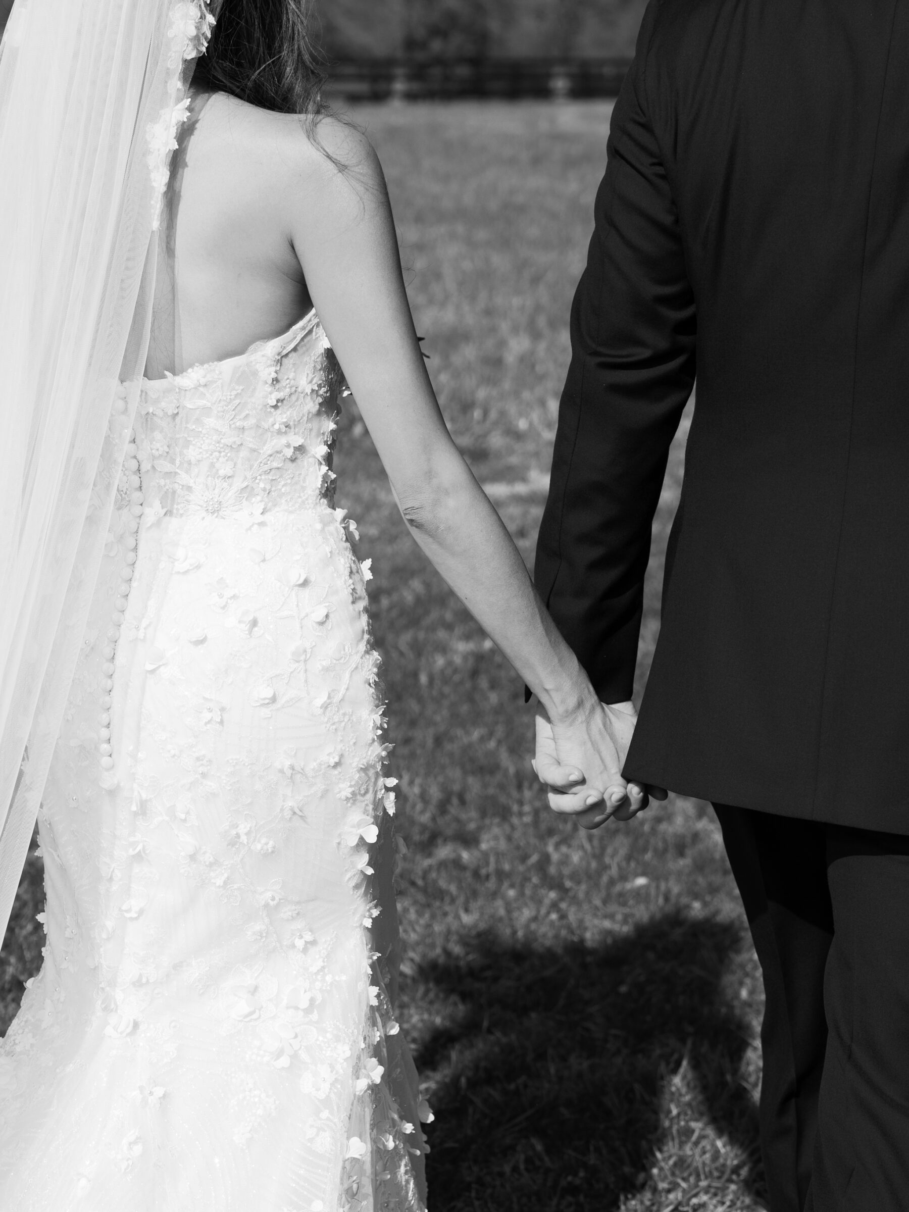 A black and white image of a bride and groom holding hands as they walk through a grassy field. The bride's intricate lace wedding dress is visible from behind, with delicate floral details on the fabric, and her veil flows gently behind her. The groom, wearing a classic tuxedo, holds her hand with a tender grip, symbolizing their connection as they walk together.