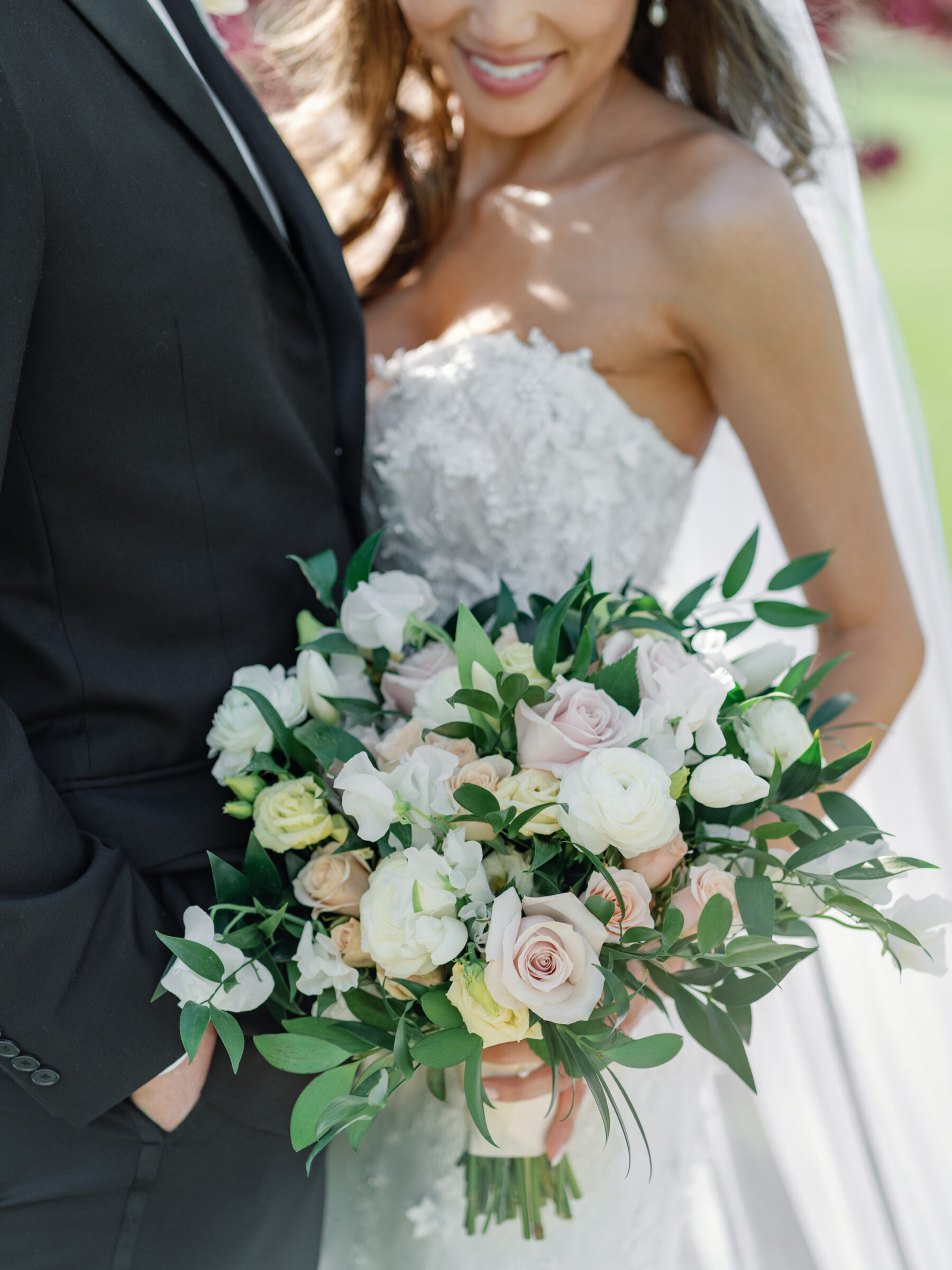 A close-up of a bride's hands holding a lush bouquet of soft pink and white roses, ranunculus, and greenery. She stands beside her groom, his hand resting on her waist, with a soft smile on her face. The vibrant colors of the bouquet contrast beautifully against her intricate wedding dress, and the blurred background hints at the outdoor setting.