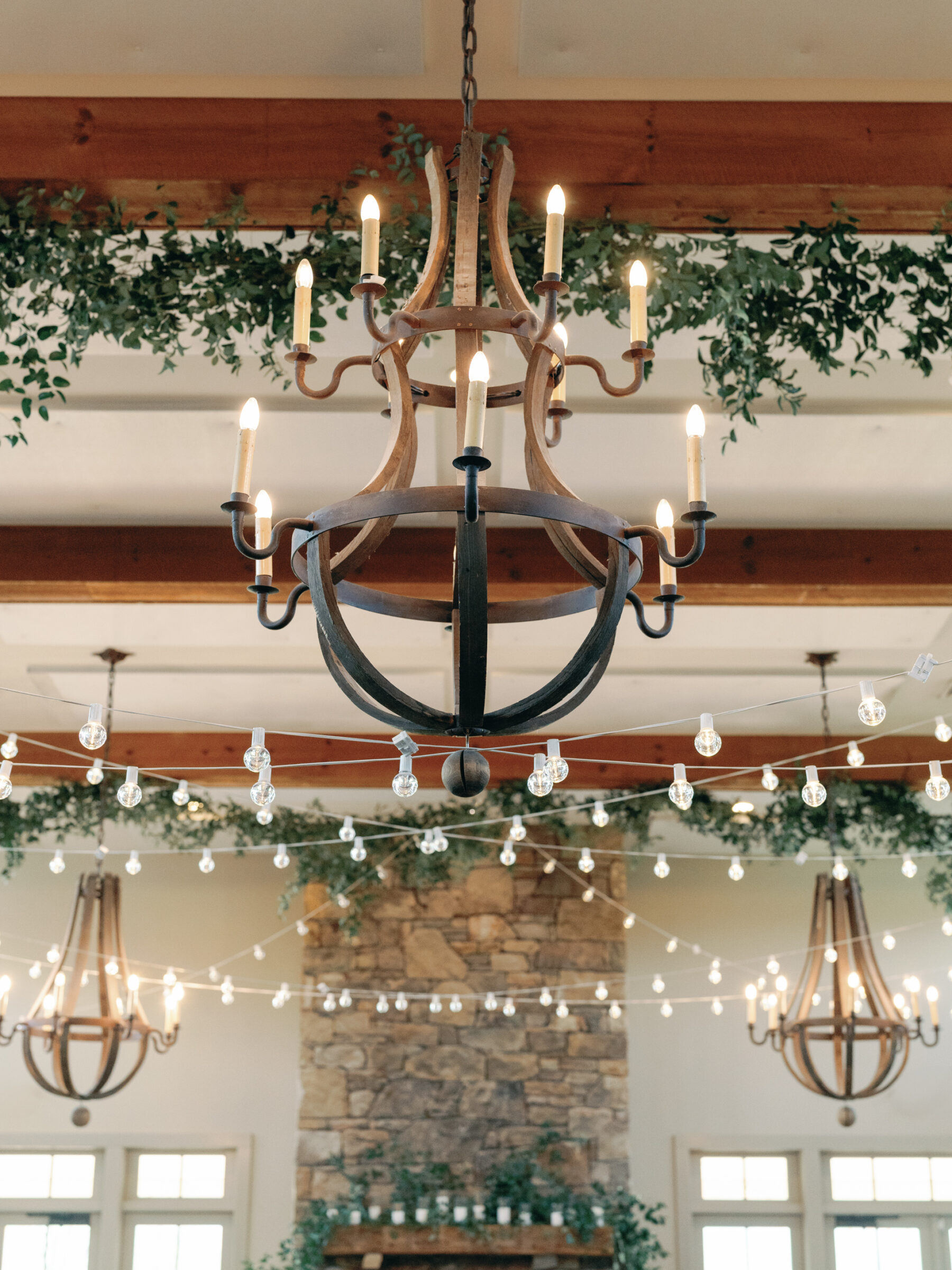 An elegant chandelier hangs from the ceiling, surrounded by strands of glowing string lights. The rustic chandeliers, with their warm, inviting light, complement the lush greenery draped along the beams above. In the background, a stone fireplace is adorned with candles, adding to the romantic atmosphere of the venue.