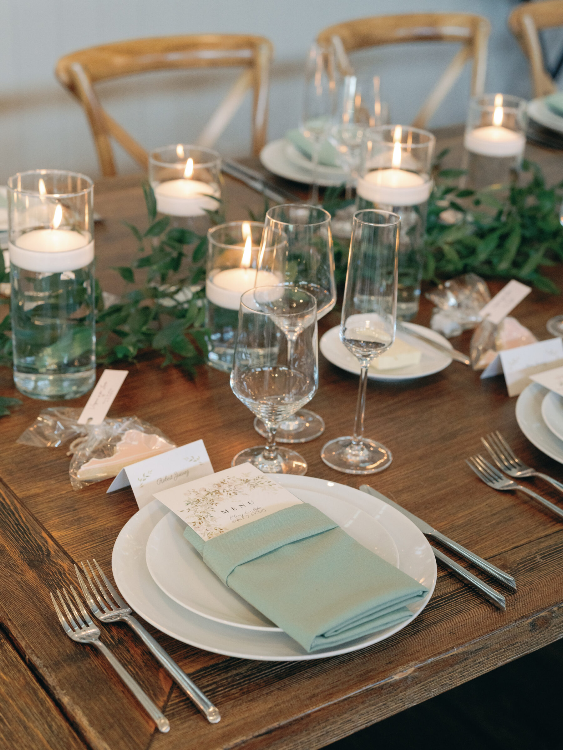 A beautifully set reception table features a soft green napkin folded neatly on white plates, with a delicate menu card atop each place setting. Tall glass vases with floating candles are surrounded by lush greenery, creating an elegant and romantic ambiance. Crystal glasses for wine and champagne are placed beside the plates, completing the refined table arrangement. Small treats and place cards add a thoughtful, personal touch to the setup.