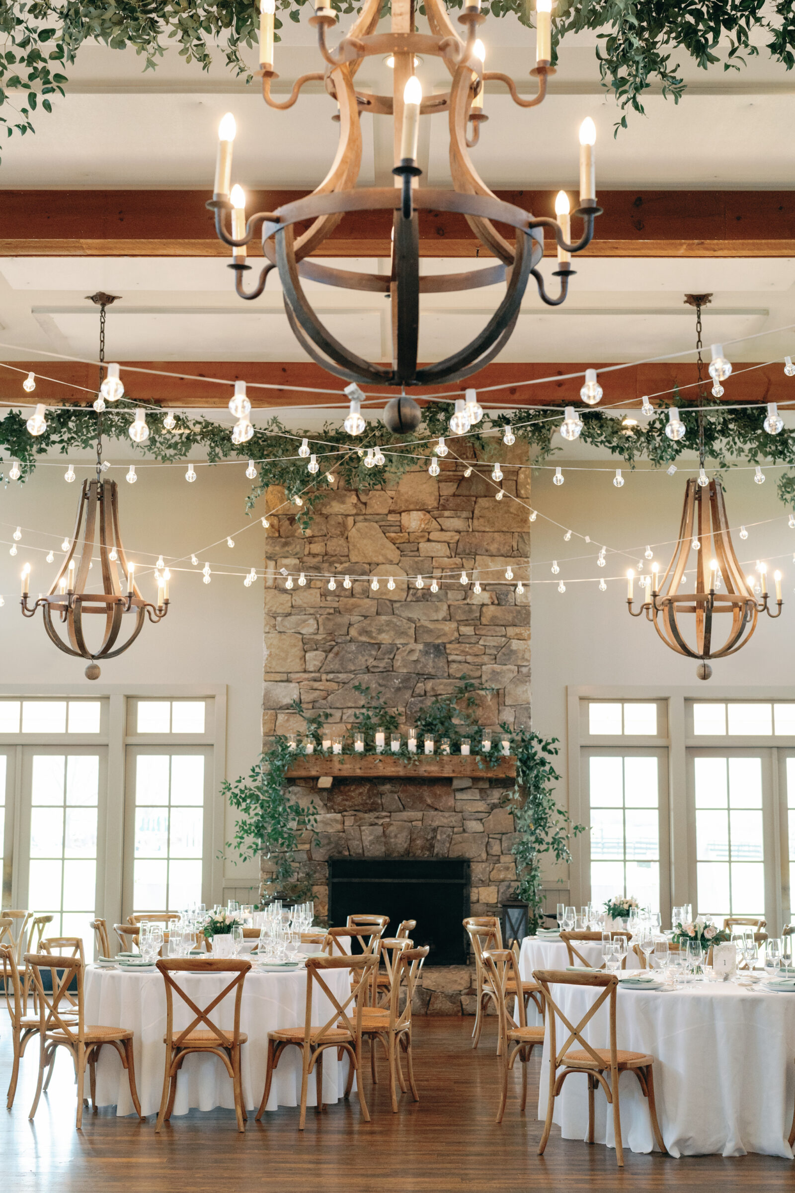 A beautifully decorated wedding reception space at King Family Vineyards. The scene features a stunning stone fireplace with candles lining the mantel, surrounded by greenery. Two rustic chandeliers hang from the ceiling, casting a warm glow over the tables below. The tables are elegantly set with white linens, clear glassware, and floral centerpieces, creating a romantic and refined atmosphere for the celebration.