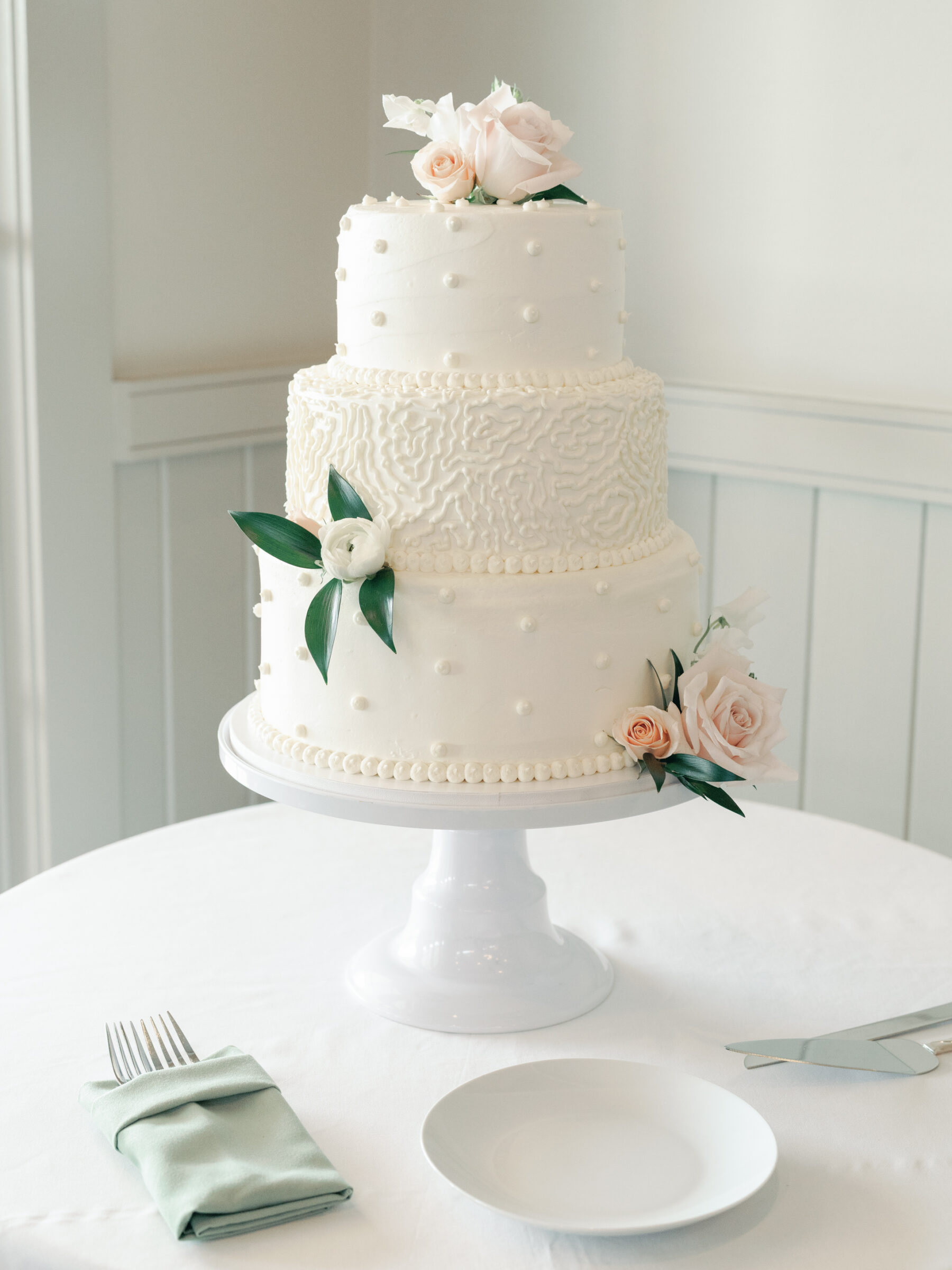 A classic three-tiered wedding cake, elegantly decorated with soft, textured white icing and adorned with delicate pearls. Fresh pale pink roses and white blooms with green leaves crown the top, adding a touch of romance. The cake sits on a white pedestal on a simple table, accompanied by a green napkin, a plate, and cake-cutting utensils, all arranged with care for a refined presentation.