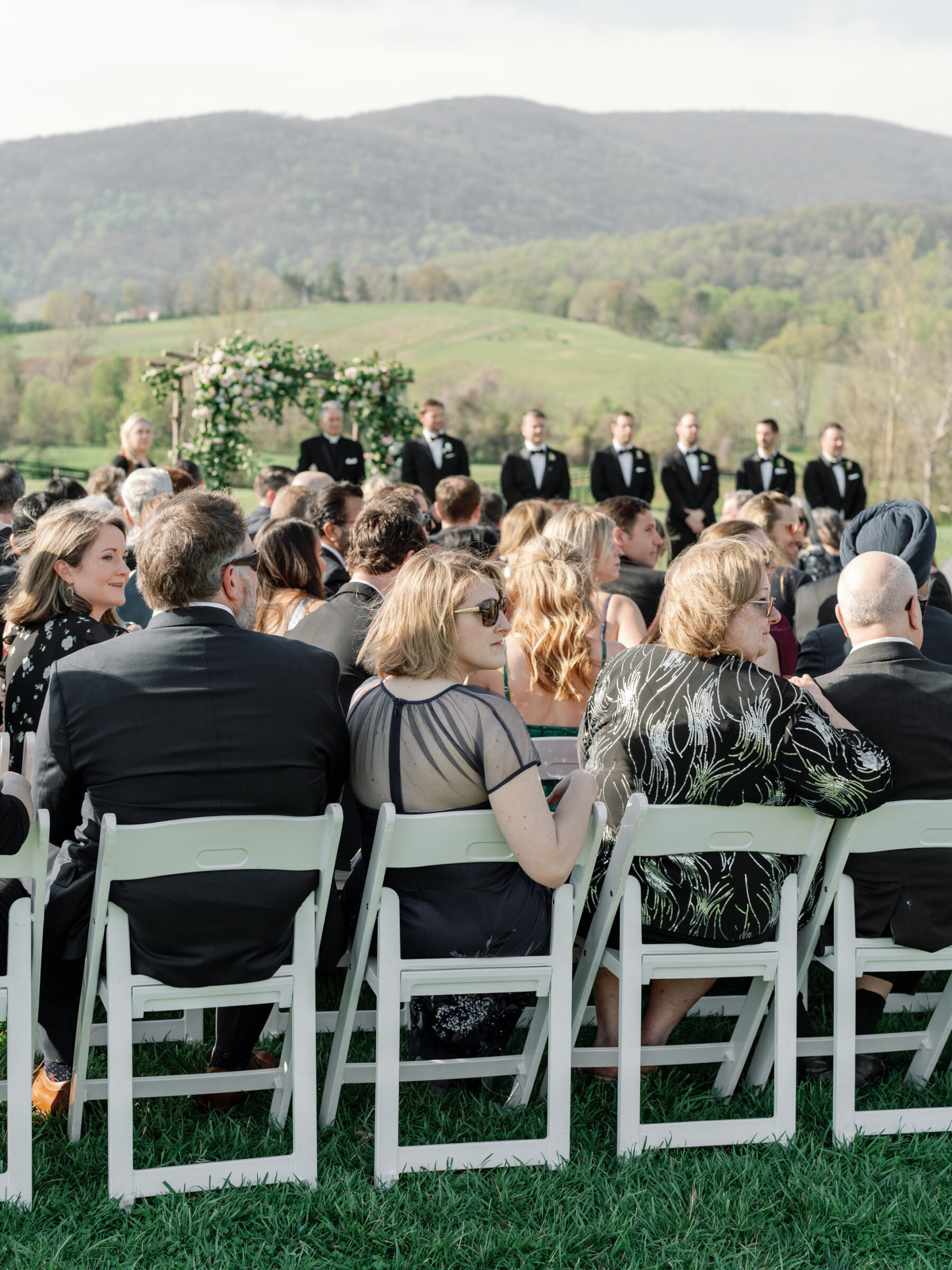 Guests sitting in white chairs at a wedding ceremony at King Family Vineyards, with the bridal party standing at the front near a flower-covered arch. The scenic mountain backdrop and lush green fields create a beautiful and serene setting for the outdoor ceremony.
