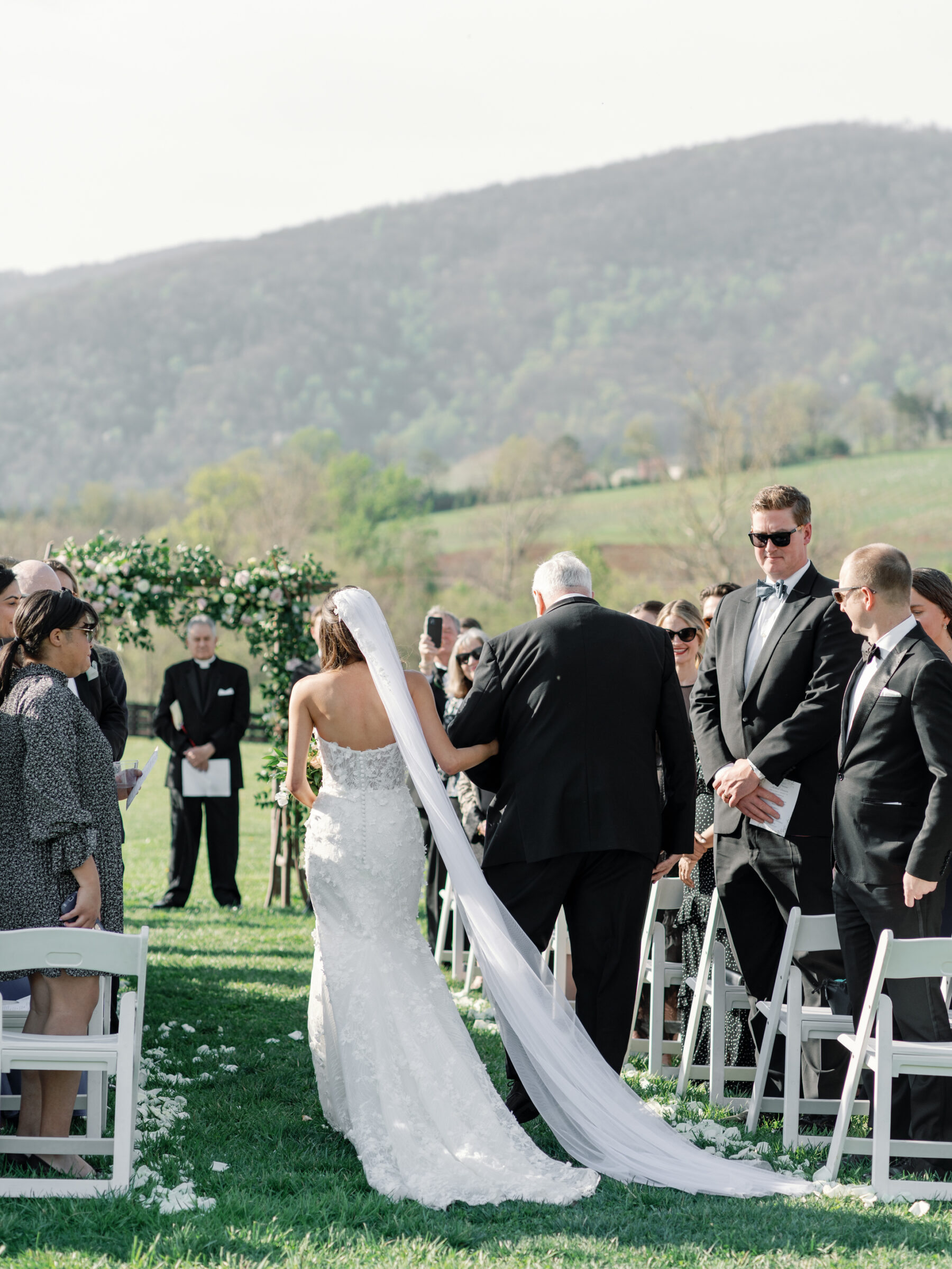 A bride walks down the aisle with her father at King Family Vineyards, with guests seated on either side. The flower-lined aisle and the beautiful mountain backdrop enhance the romantic atmosphere of the outdoor ceremony.