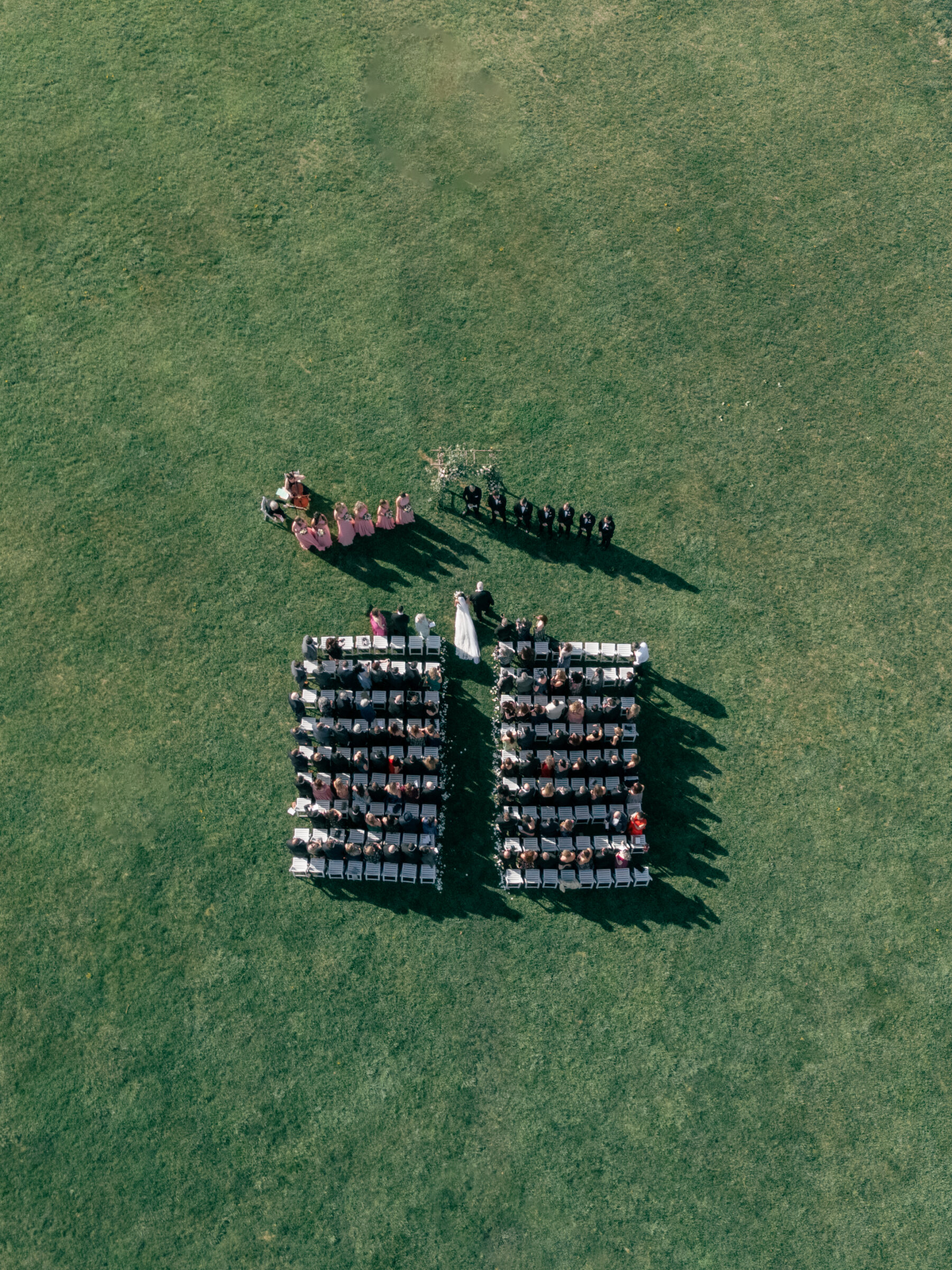 Aerial view of the wedding ceremony at King Family Vineyards, with guests seated in neat rows and the wedding party standing at the altar. The expansive green lawn and mountainous backdrop create a stunning, serene setting.