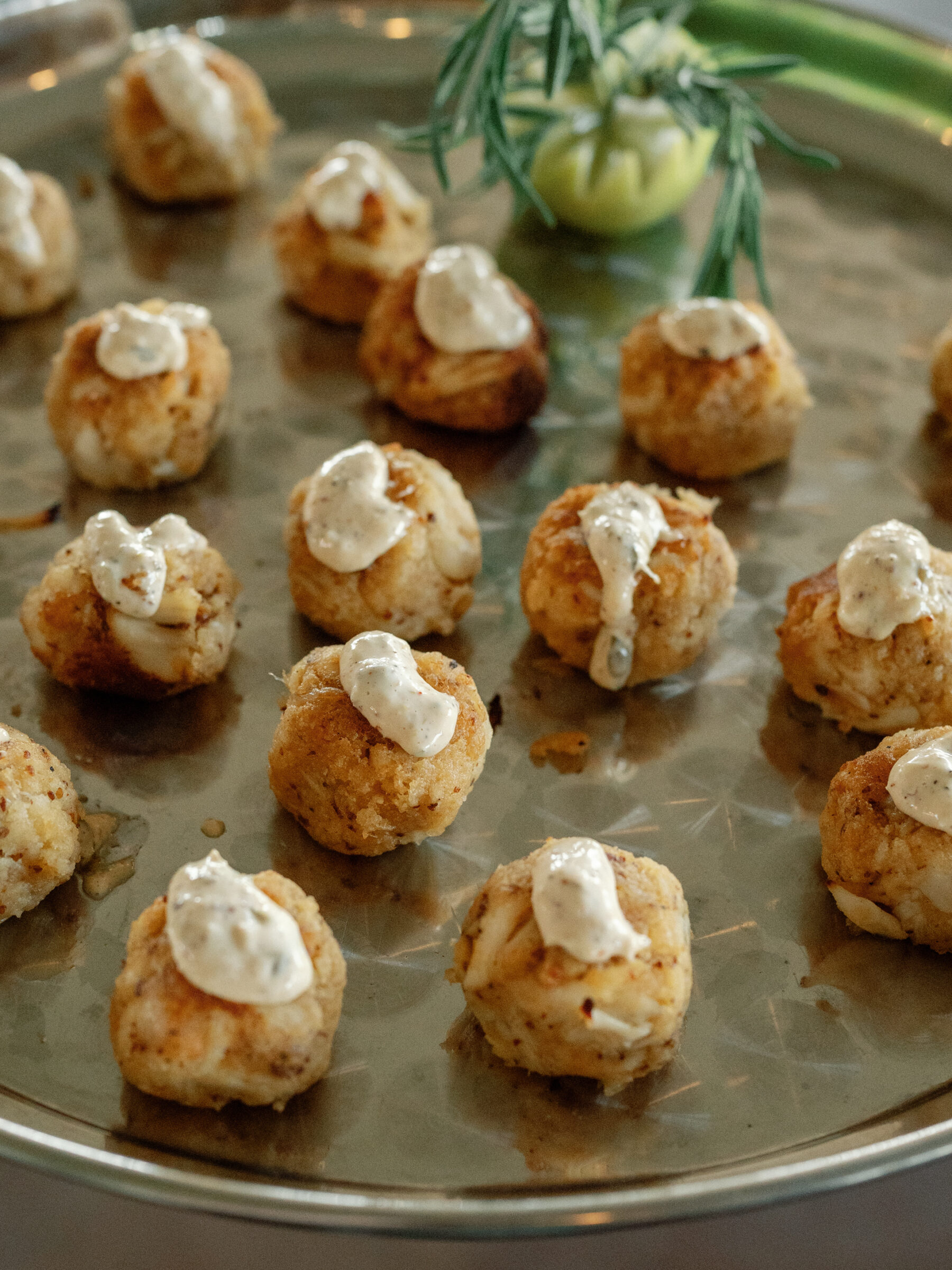 A close-up shot of crab cakes topped with a dollop of creamy sauce, served on a silver tray with a sprig of rosemary in the background.