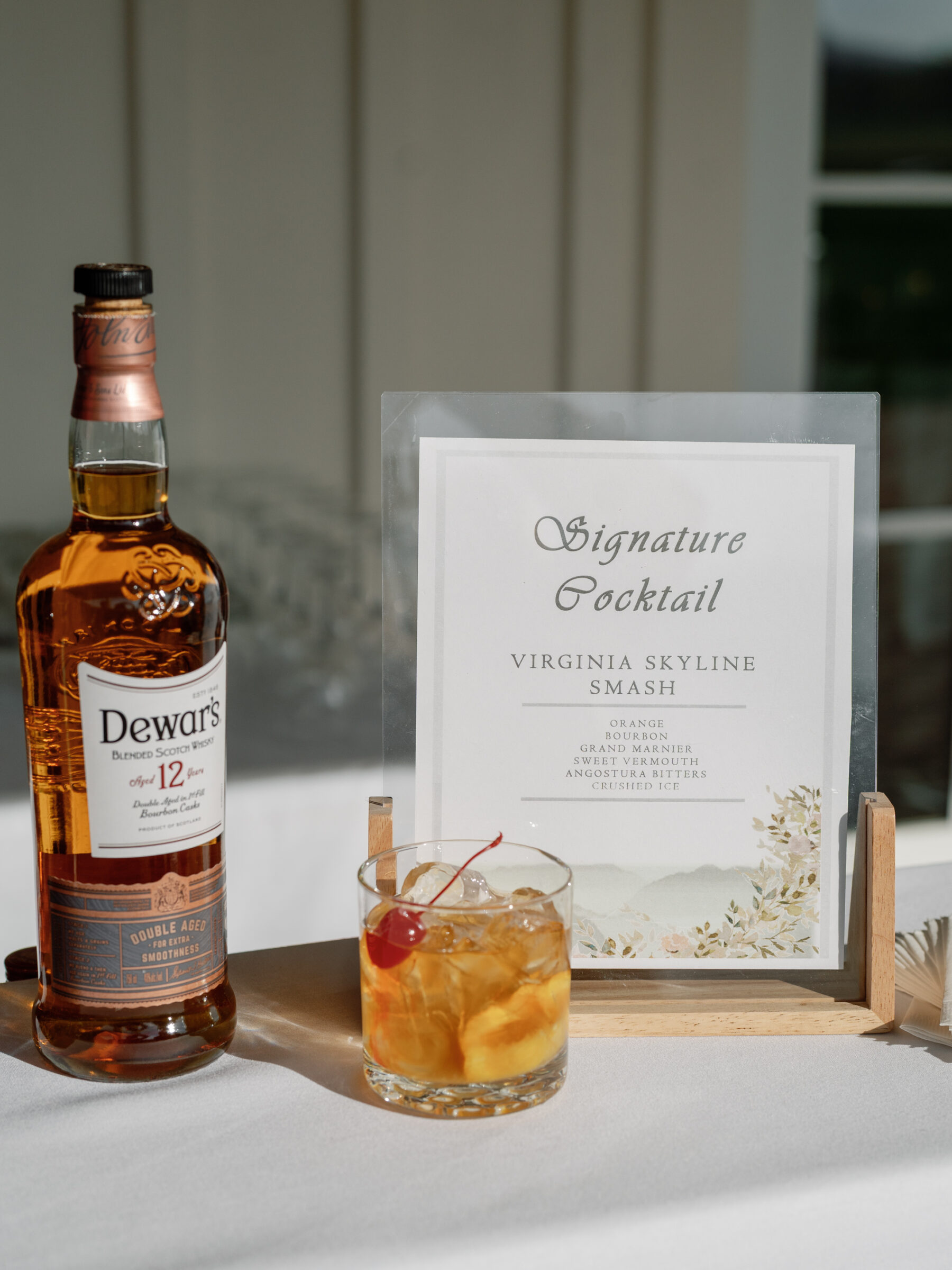 A bottle of Dewar's 12-year-old blended Scotch whiskey stands next to a glass of the "Virginia Skyline Smash" signature cocktail, with a cherry on top, accompanied by a printed menu card displaying the cocktail's ingredients.