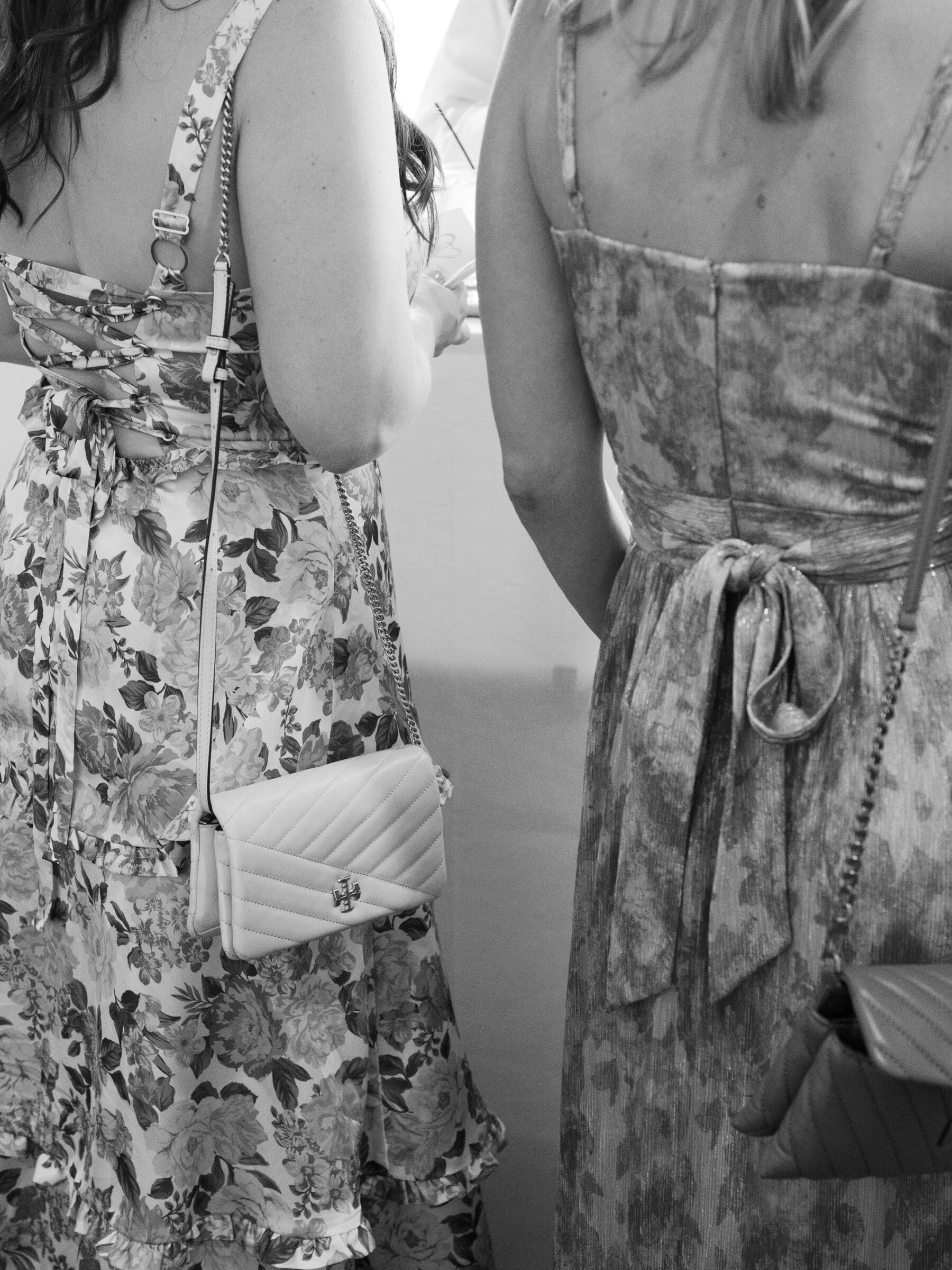 A black-and-white photograph of two women in floral dresses standing together, holding small handbags, at a wedding event.