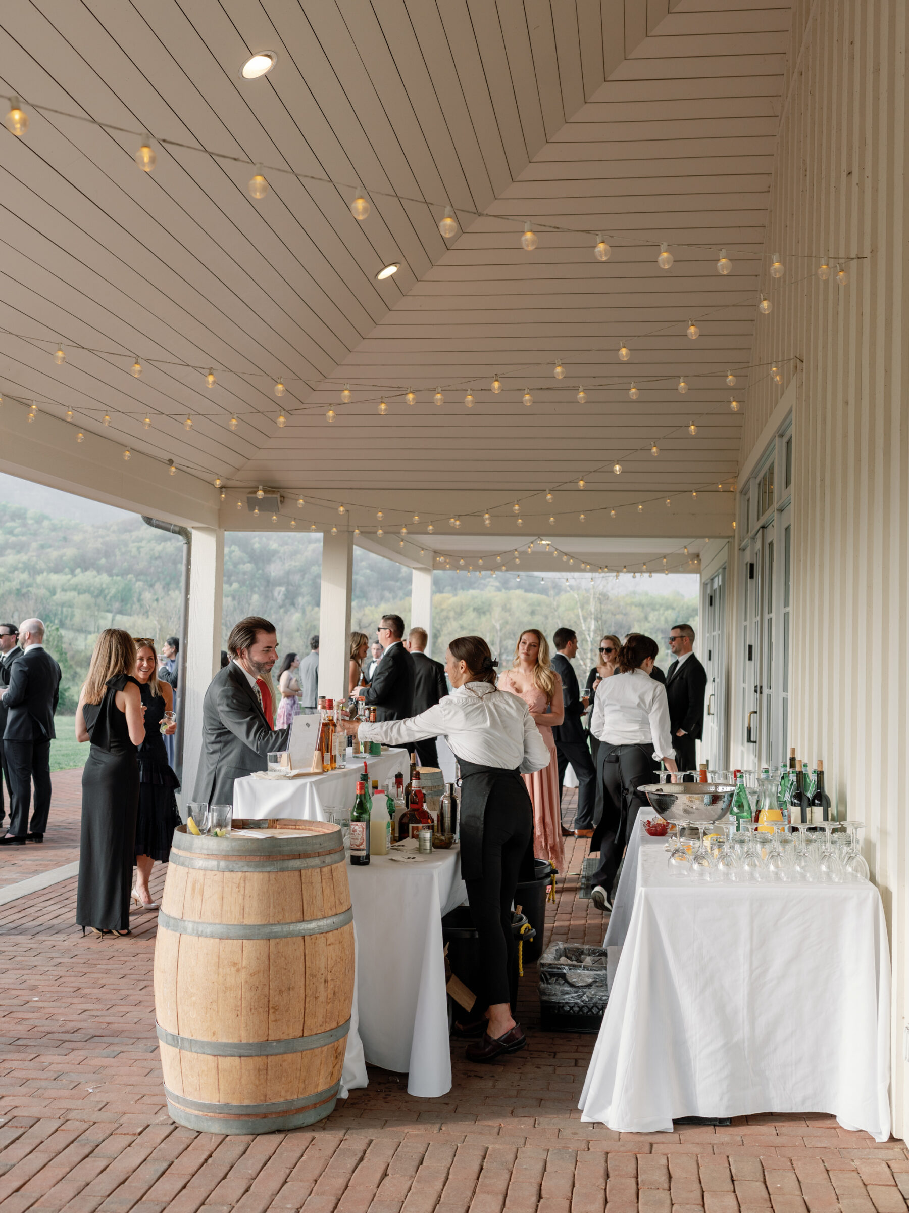 A beautifully lit outdoor bar area during cocktail hour at a wedding, with guests enjoying drinks and a bartender serving at a rustic barrel table.