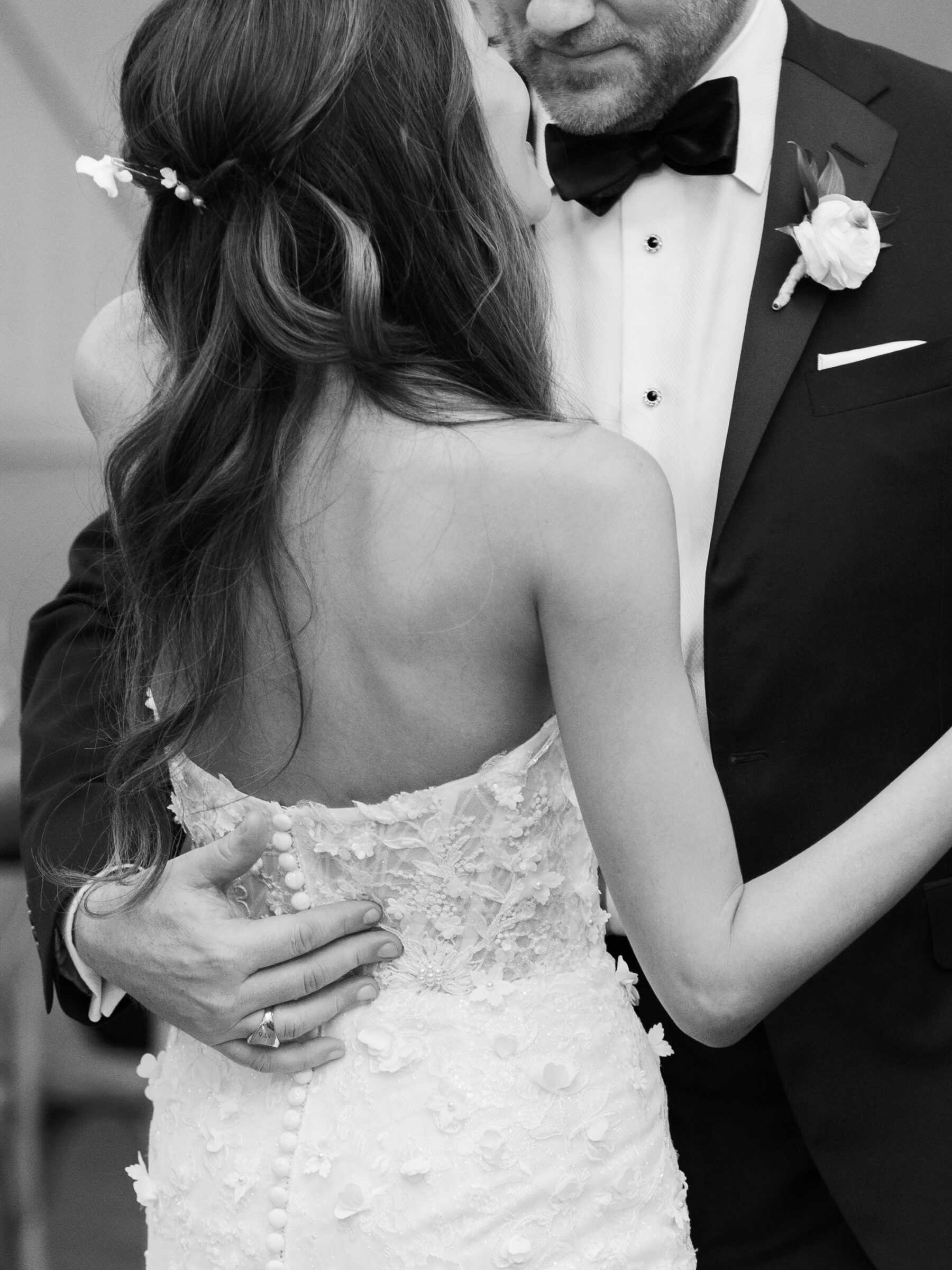 A close-up of the couple during their first dance, with the bride’s back and gown delicately displayed as she rests her hand on her groom’s chest. His hand gently holds her waist. The intimate moment is enhanced by the soft lighting, and the bride’s hair is adorned with small white flowers, adding to the elegance of the scene.
