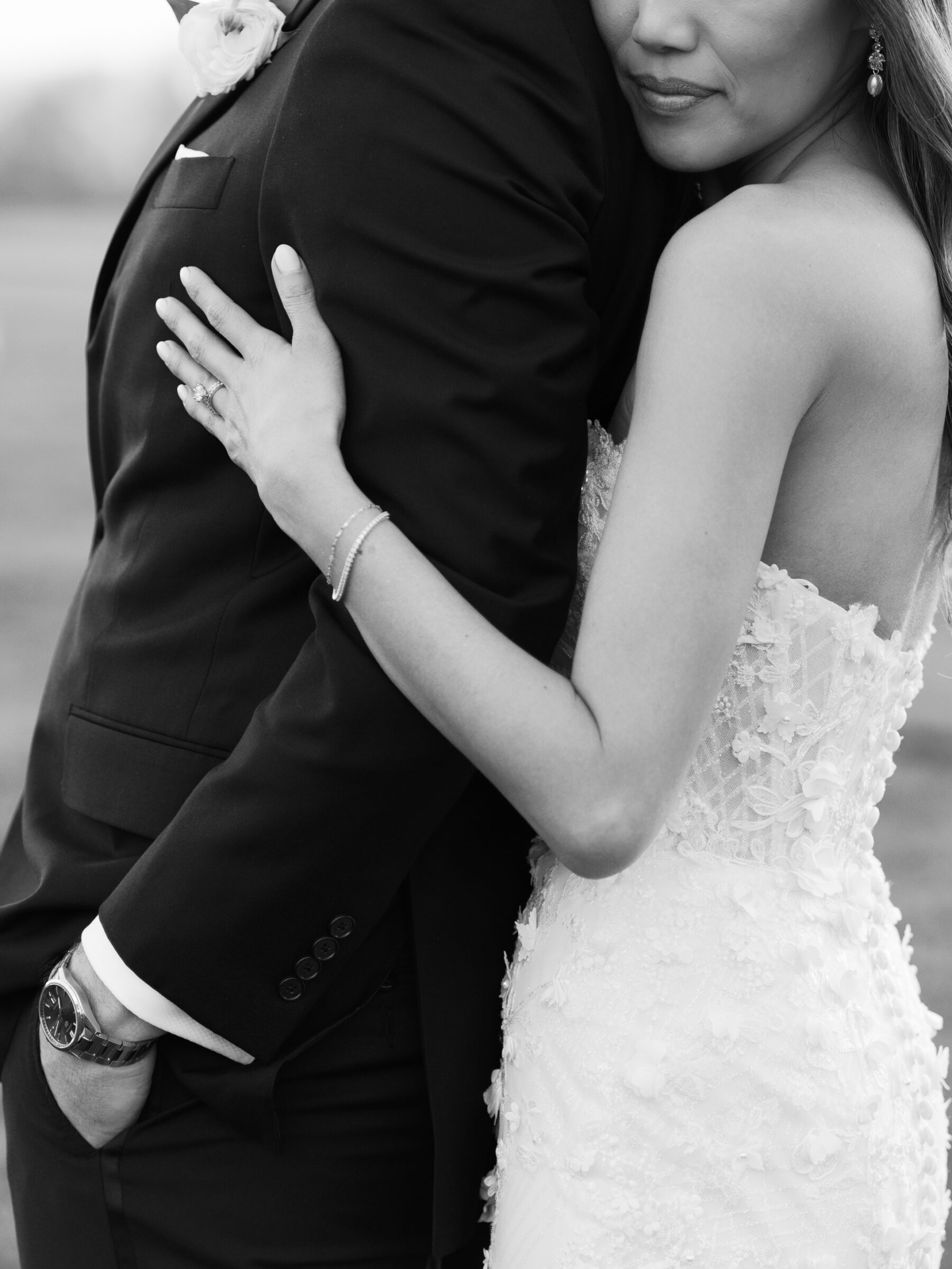 A black and white close-up of a couple in a tender embrace. The bride, dressed in a detailed lace gown, holds her groom’s arm, while her other hand rests gently on his chest. The groom, in a classic black tuxedo, gazes down at her, his arm wrapped around her waist. The focus is on the emotional connection between them, with subtle details of the wedding attire highlighted in the soft light.